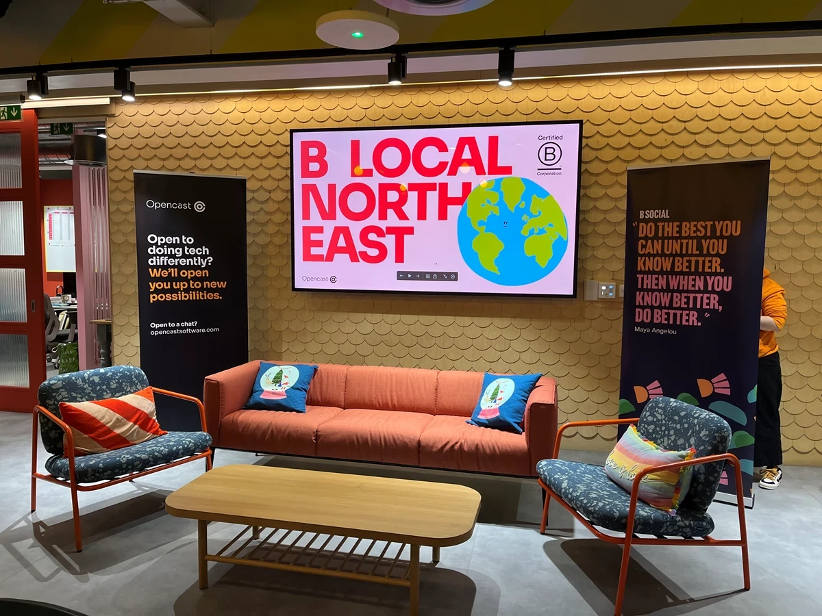 Modern seating area with a red sofa, two chairs, and a screen reading “B Local North East” with an Earth graphic.
