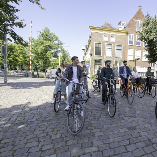 En grupp människor cyklar på en kullerstensgata med träd och byggnader i bakgrunden under en klar himmel.