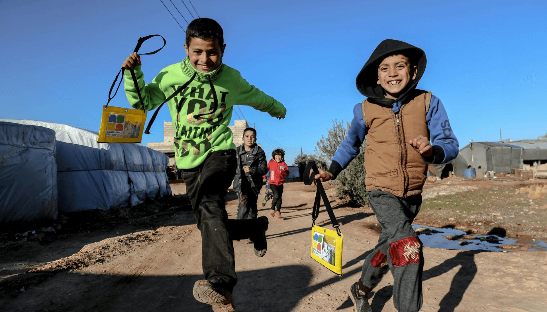 Three smiling children stand in a grassy area in front of a light-colored building, wearing colorful jackets. Behind them, stacked white cardboard boxes display the phrase "REBUILD THROUGH PLAY."