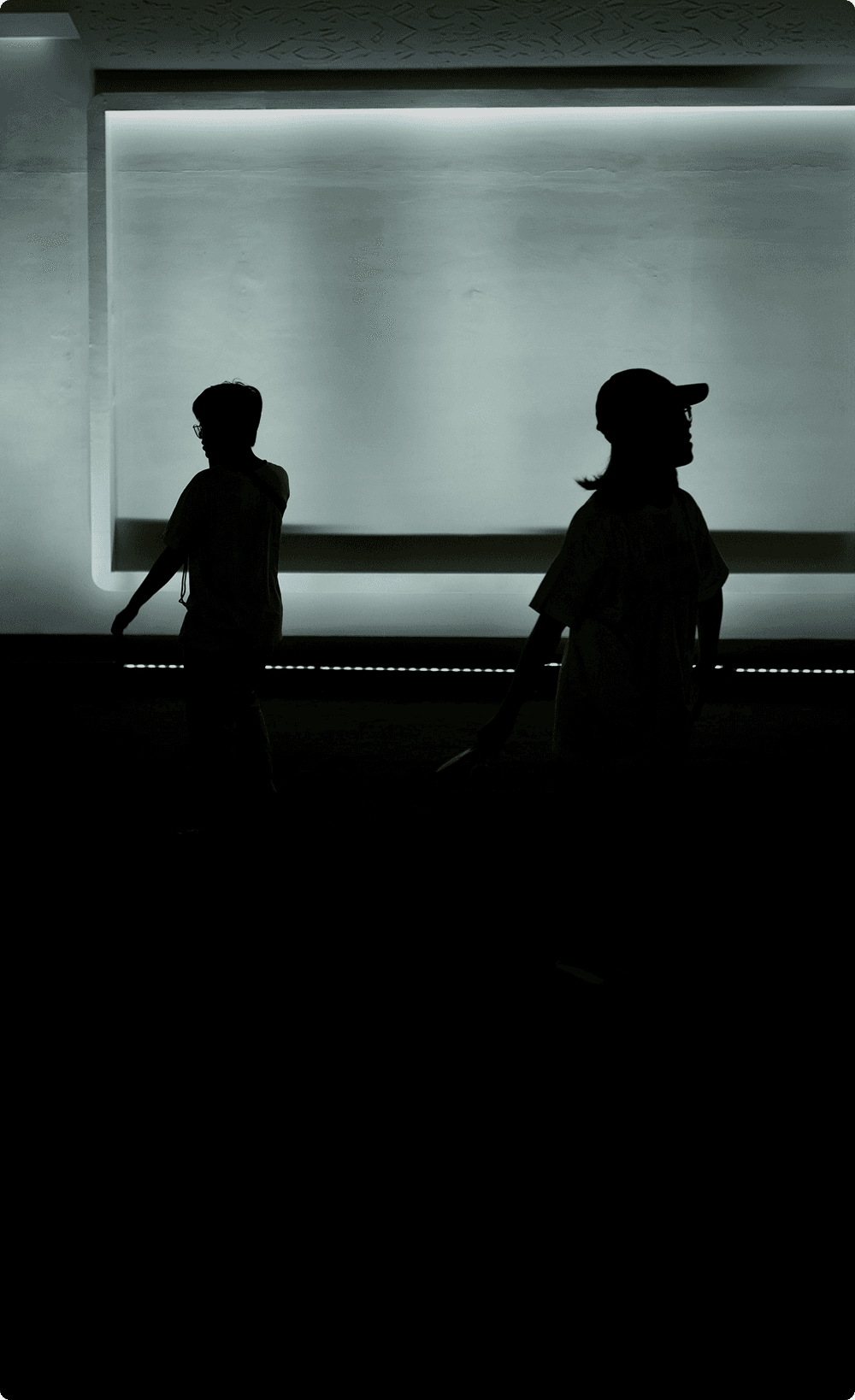A silhouette of two people walking past a large, brightly glowing rectangular light fixture on a dark wall.