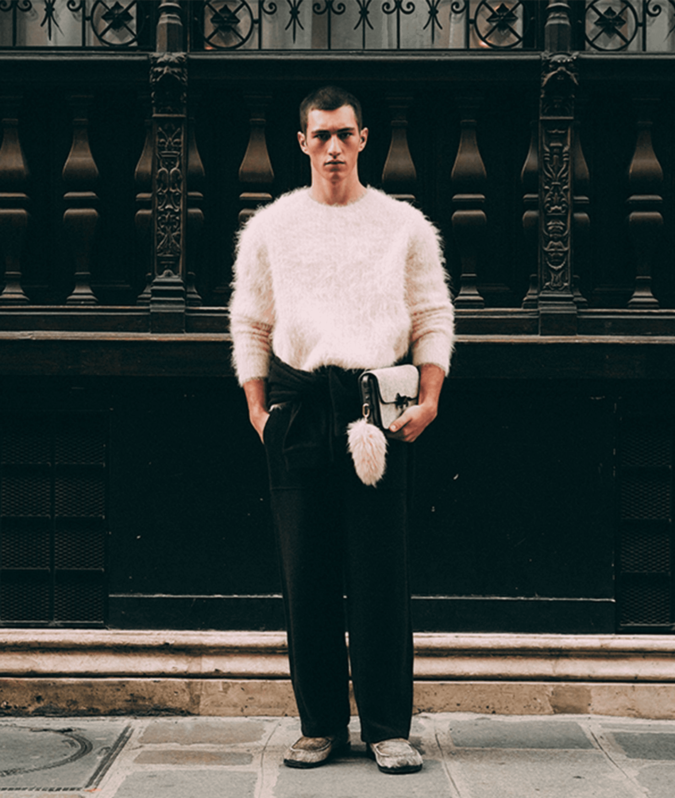 A person with shoulder-length hair is wearing a tiger print sweater and high-waisted black leather shorts, holding a black clutch with a gold accent, standing on sunlit stone steps.
