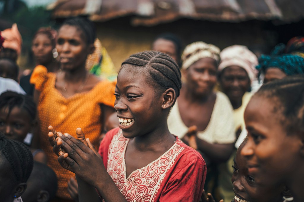 A young girl claps and smiles joyfully in the midst of a group of women and children, celebrating together in a vibrant, close-knit community gathering.