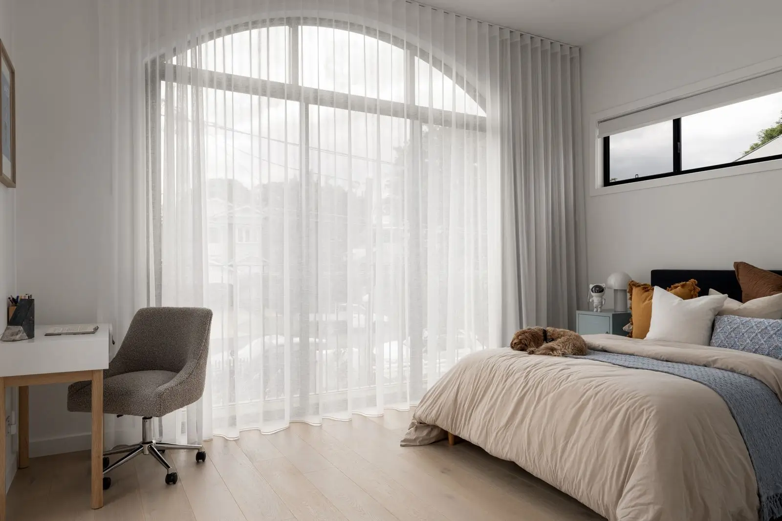 A bedroom with sheer curtains and a desk and chair in the far left hand side