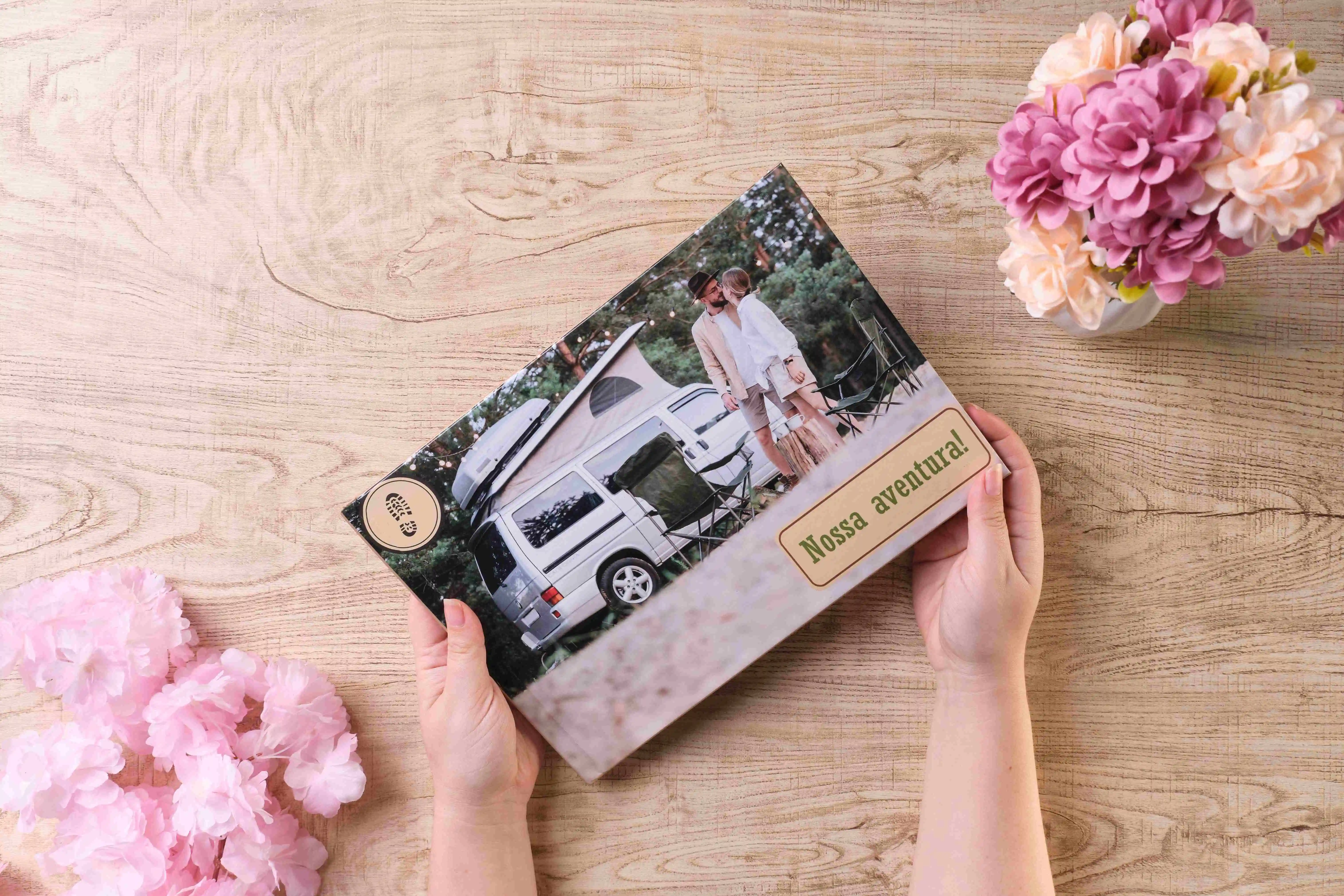 Pessoa segurando fotolivro personalizado com capa de casal em frente a uma van de viagem, título ‘Nossa aventura!’, sobre mesa de madeira decorada com flores.