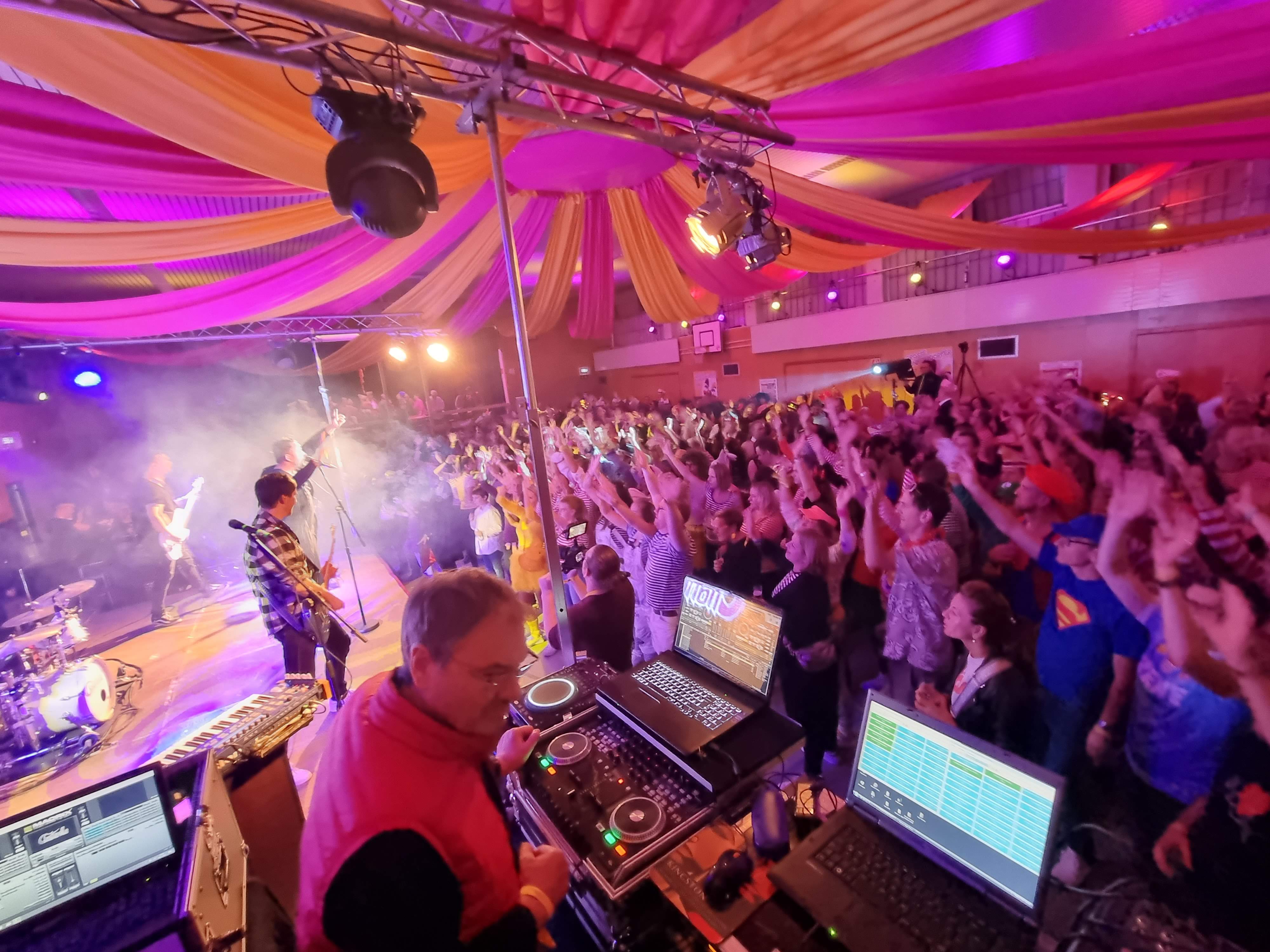 Mann, DJ, der am Mischpult steht und gute Laune und Stimmung in einer Halle verbreitet. Dabei wird eigene Licht- und Tontechnik verwendet, die auf dem Bild sichtbar ist.