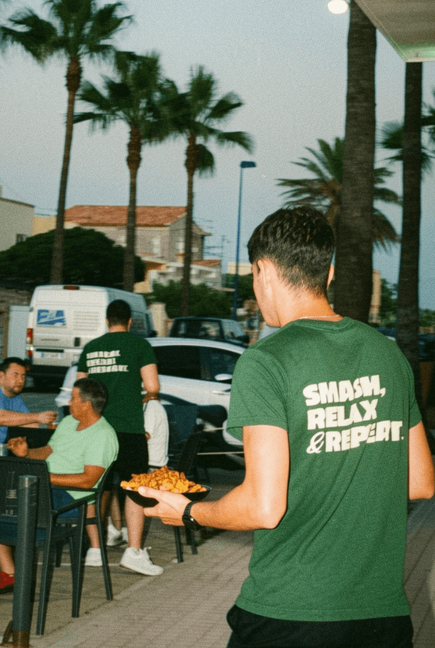 Person with a mustache wearing a black t-shirt carrying a tall stack of pizza boxes inside a dimly lit bar or restaurant.