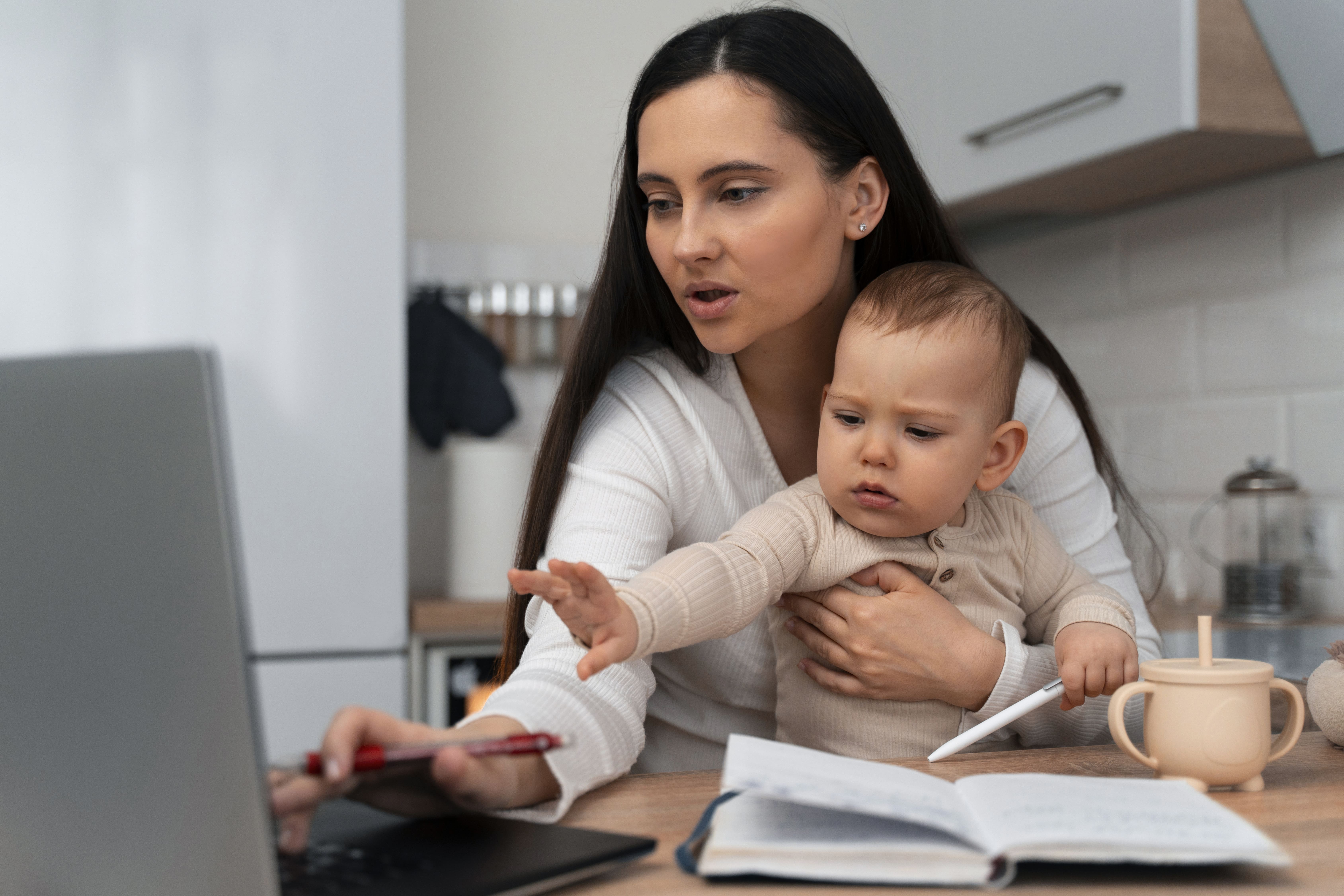 Mulher segurando um bebê enquanto trabalha em frente ao laptop. Ela está concentrada na tela, enquanto o bebê, no colo dela, estende a mão, parecendo interessado no computador. Na mesa, há um caderno aberto, uma caneca infantil e uma caneta. A cena mostra o desafio de conciliar maternidade e trabalho.