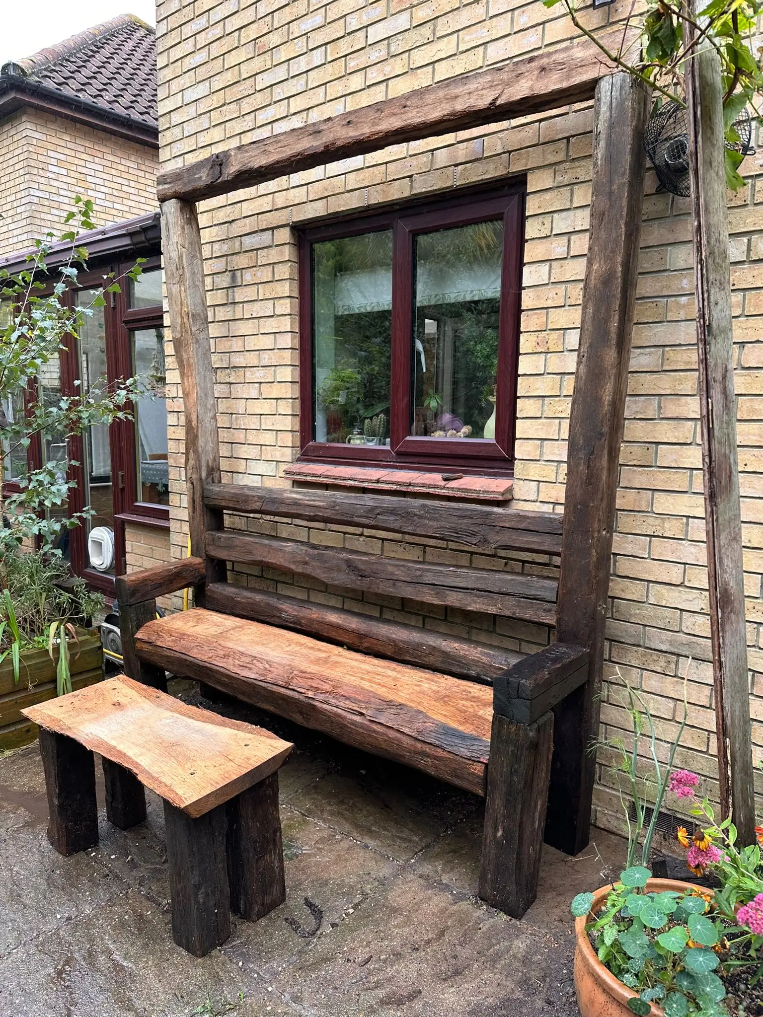 A wooden bench and small table placed beside a house, surrounded by greenery and a brick wall.