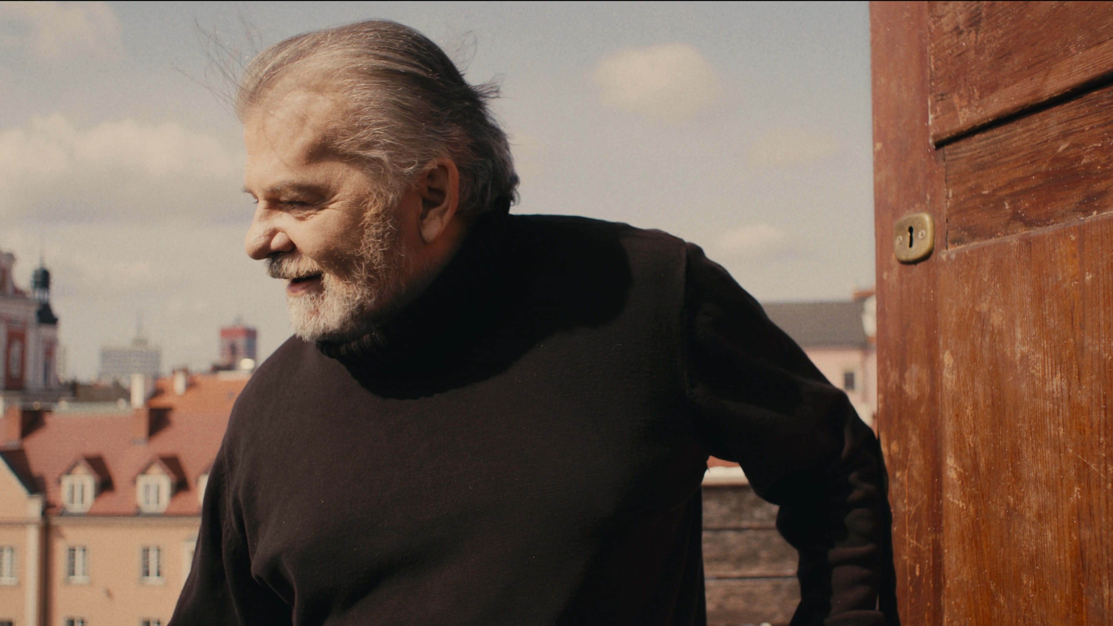 Medium shot of a man in a black turtleneck standing outdoors near a brick wall.