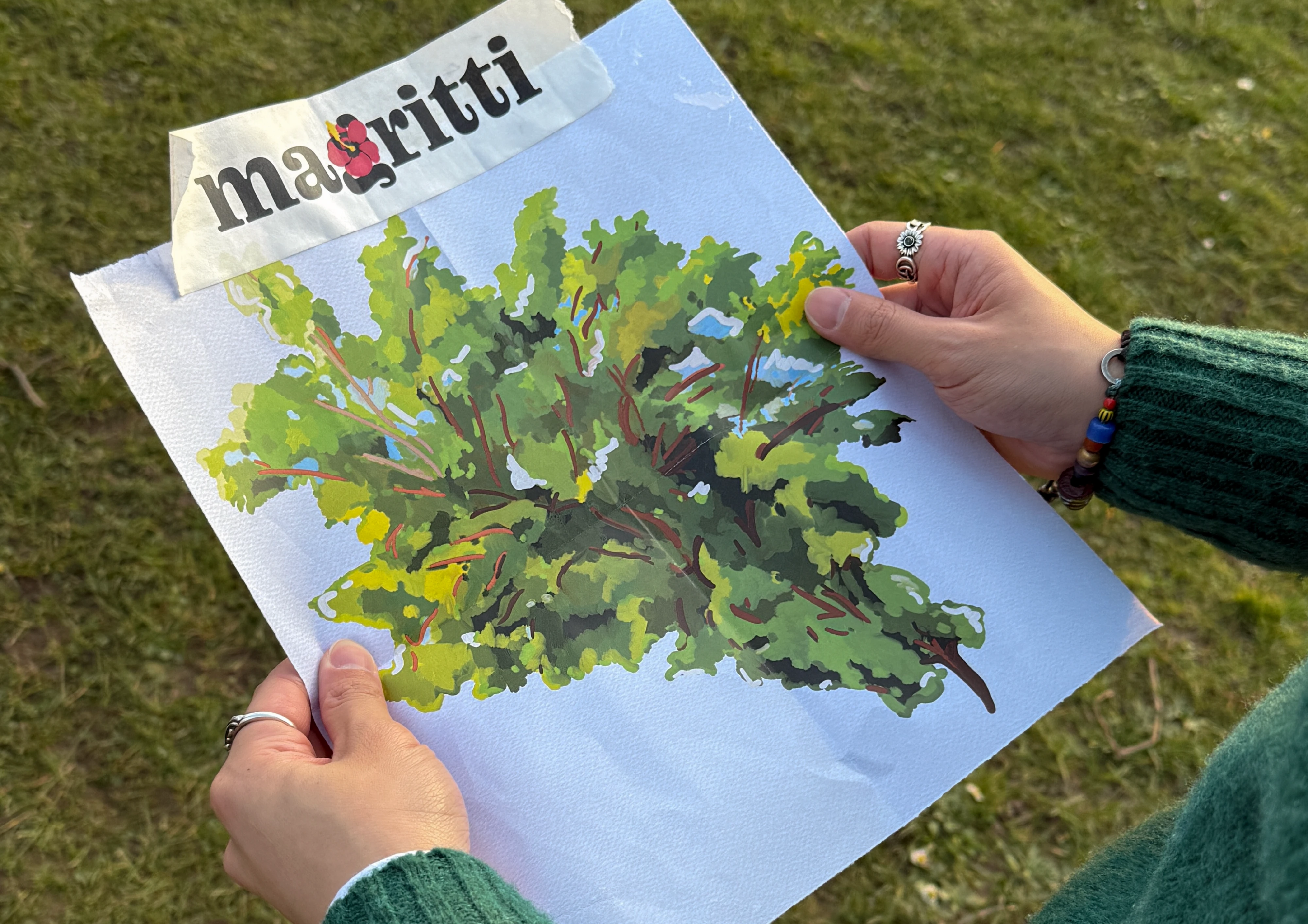 Two hands hold a painted print of a leafy green shrub above grass. A torn paper strip bearing the Magritti wordmark, its g replaced by a red hibiscus, is taped across the top edge.