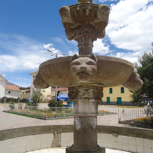 石頭噴泉，雕刻有動物面孔，位於鋪砌的廣場上，周圍有建築物和綠色植被，天空部分雲遮。