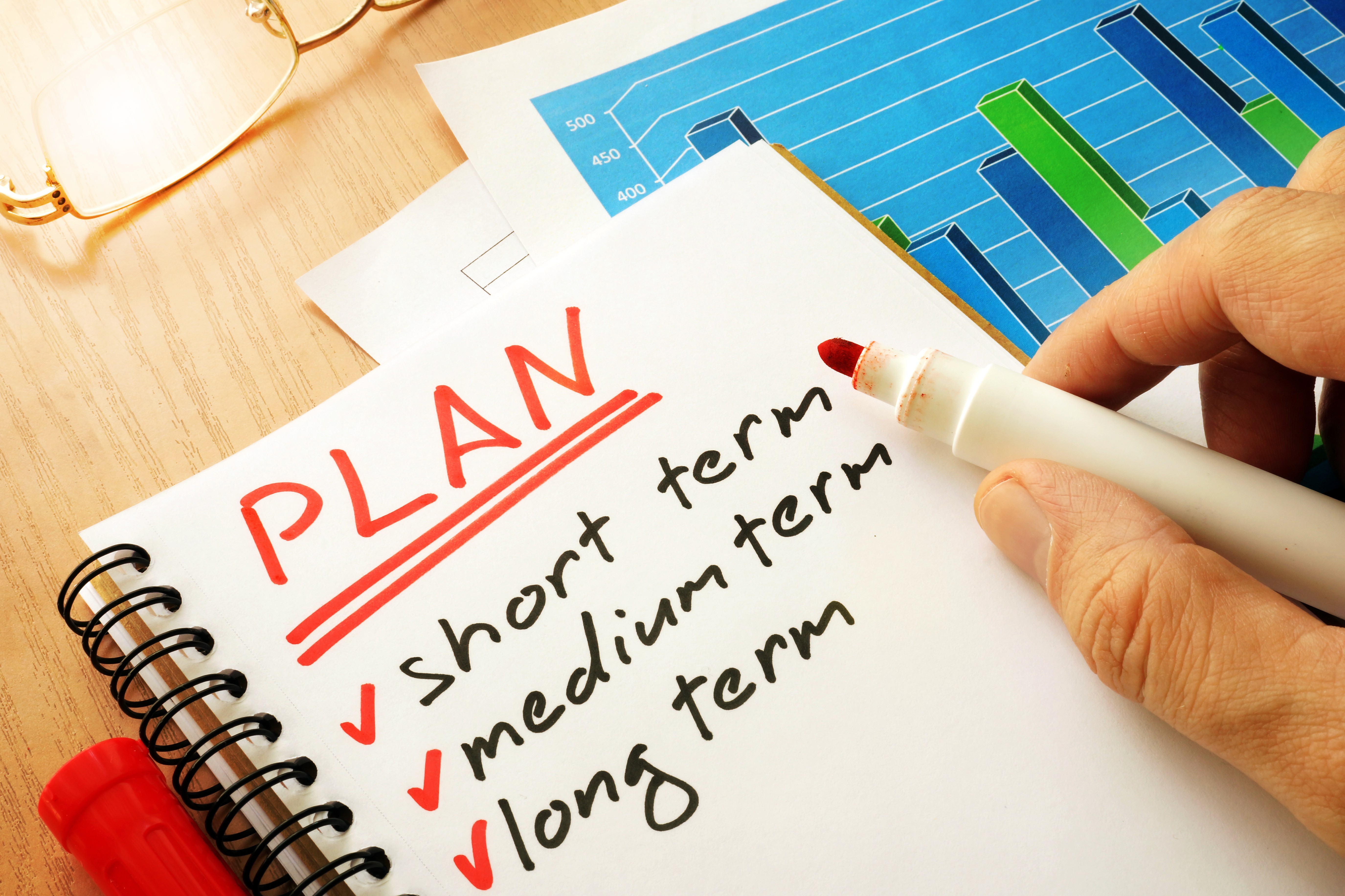 Hand writing a financial plan with short, medium, and long-term goals on a notepad beside charts and documents.