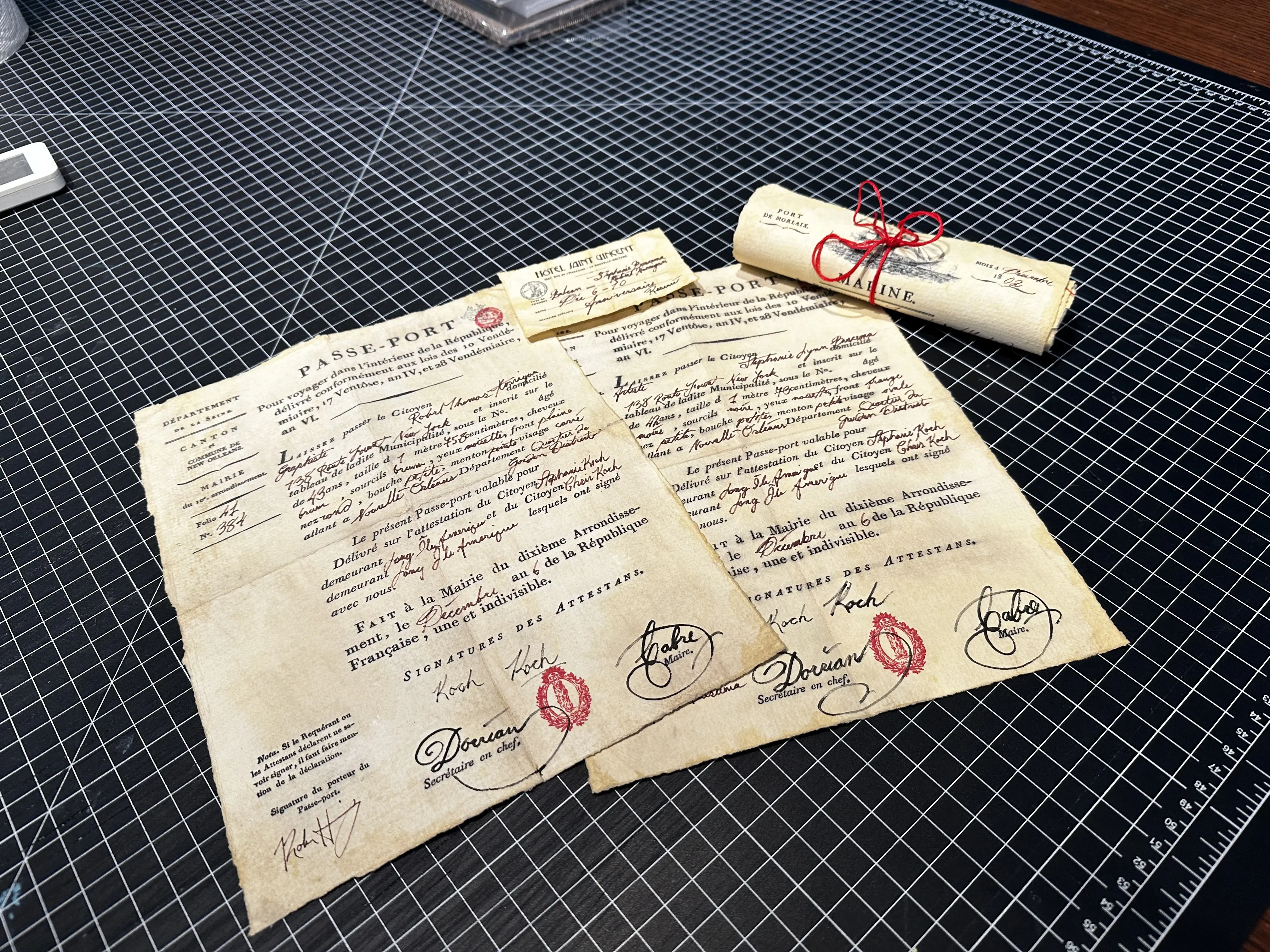 A collection of vintage-style documents lies on a grid-lined cutting mat, featuring ornate handwriting, red wax seals, and signatures, alongside a scroll tied with a red ribbon, exemplifying intricate design elements reminiscent of Rob Harrigan's work.