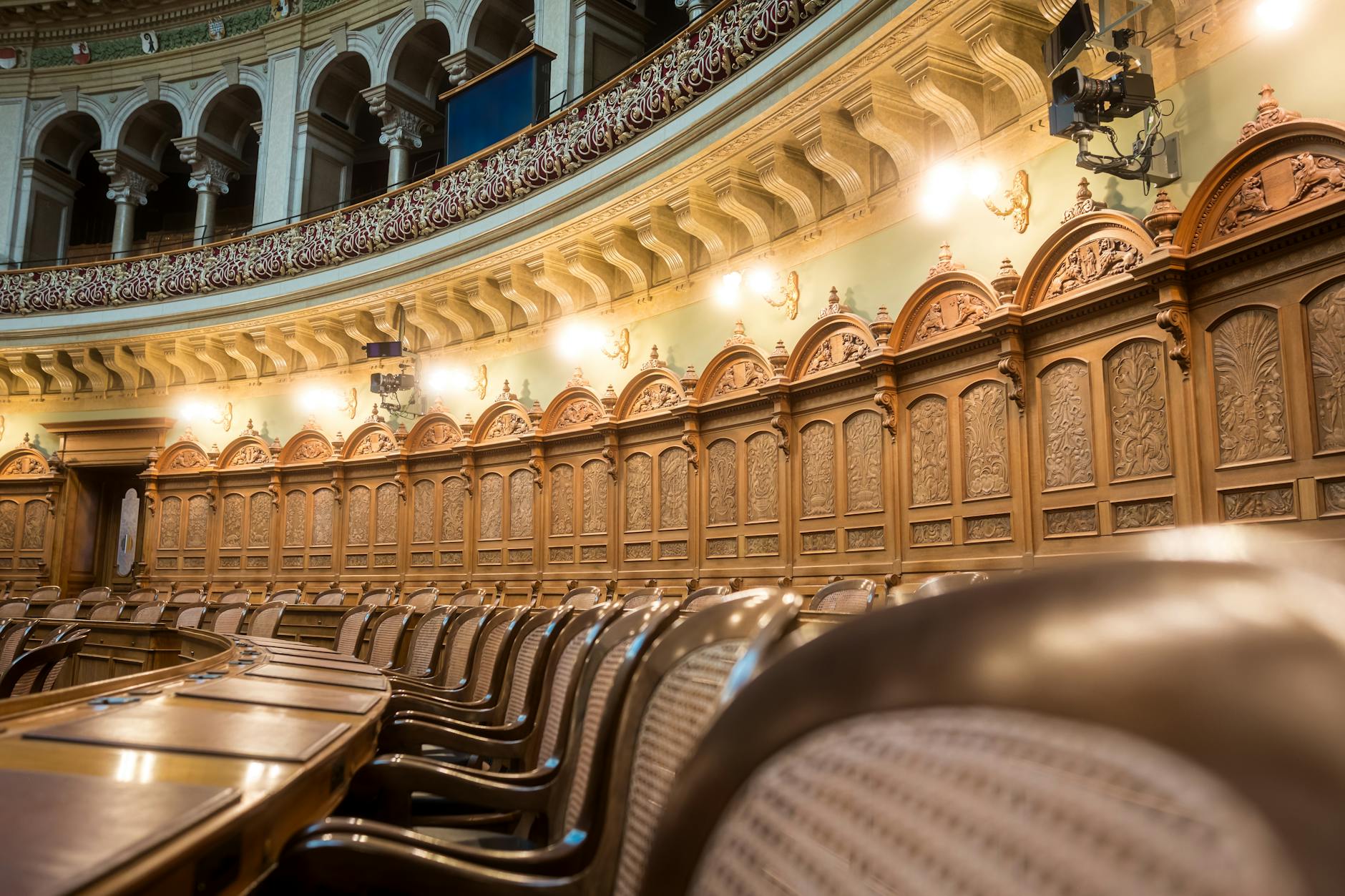 Ornate parliamentary chamber interior with rows of carved wooden seats under an arched gallery and warm hanging lamps.