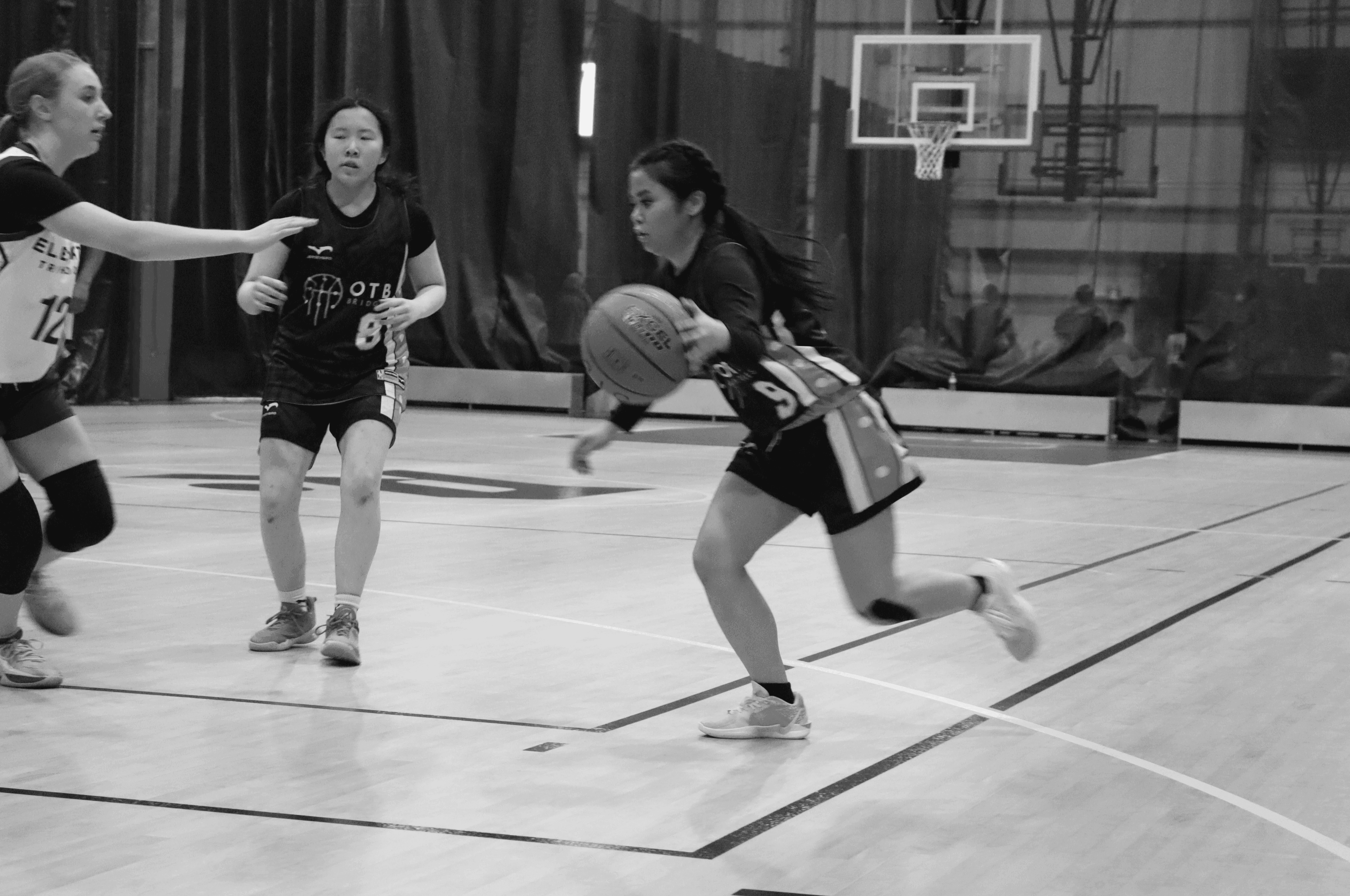 A youth basketball player in an orange jersey dribbling aggressively past a defender during a game, with a referee and spectators visible in the background on an indoor court.