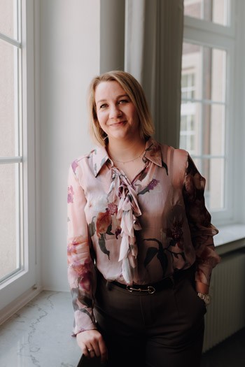 Jüngere Geschäftsführerin mit blumiger Bluse steht vor einem Fenster im Büro von SLB und blickt in die Kamera.