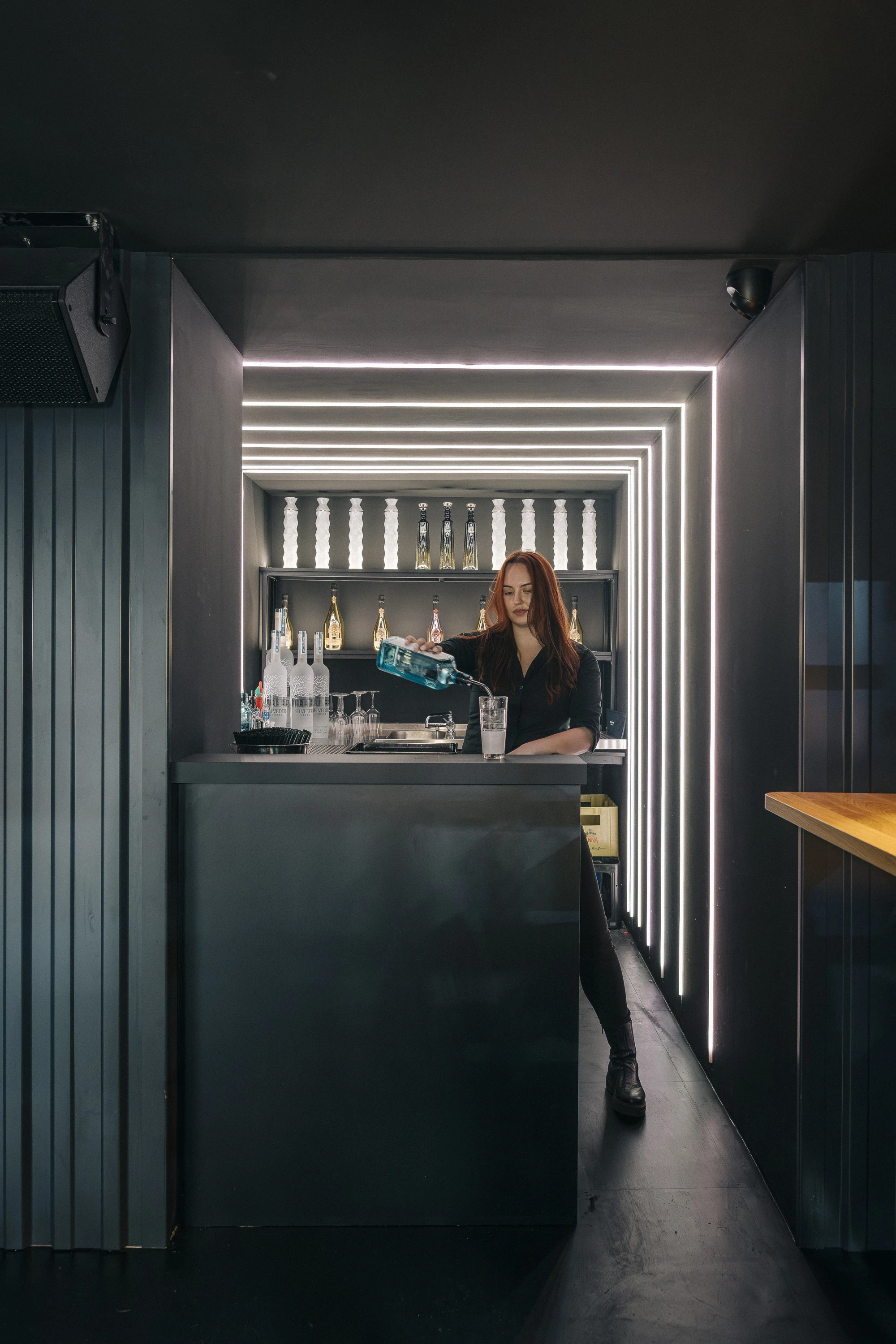 Narrow minibar niche at NATE CLUB in black, framed by horizontal white LED lines, with a bartender pouring a drink at the dark counter.