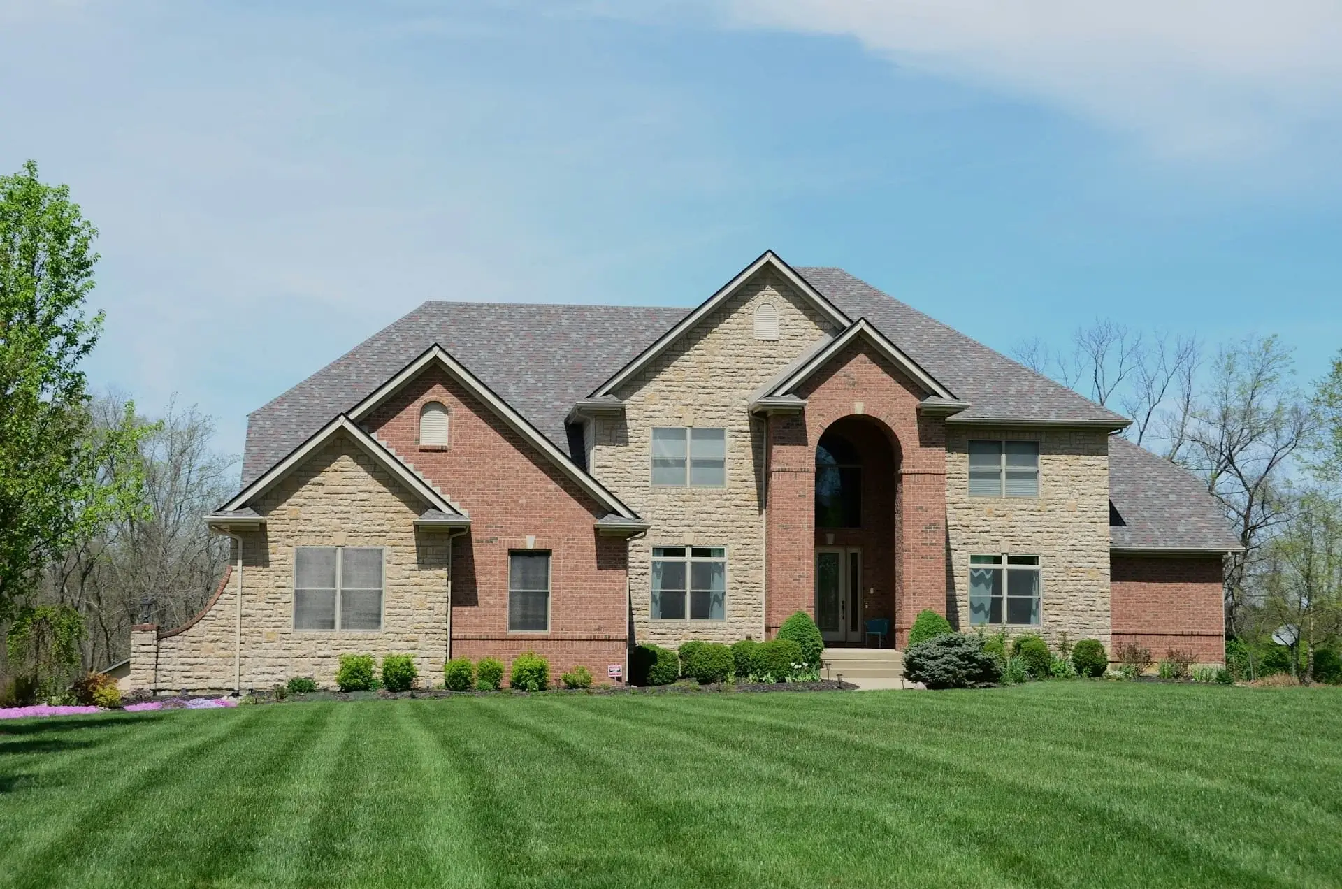 Large luxury house with a stone and brick exterior, arched entrance, symmetrical windows, and a wide green front lawn.