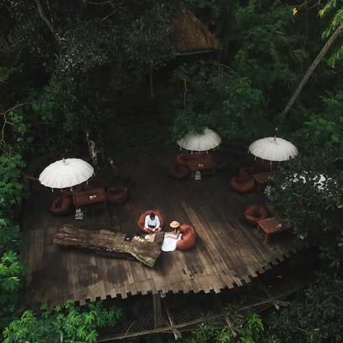 無人機視角下的森林中木平台，擺放著桌子、椅子、白色陽傘，和兩個人坐在一張大木桌旁。