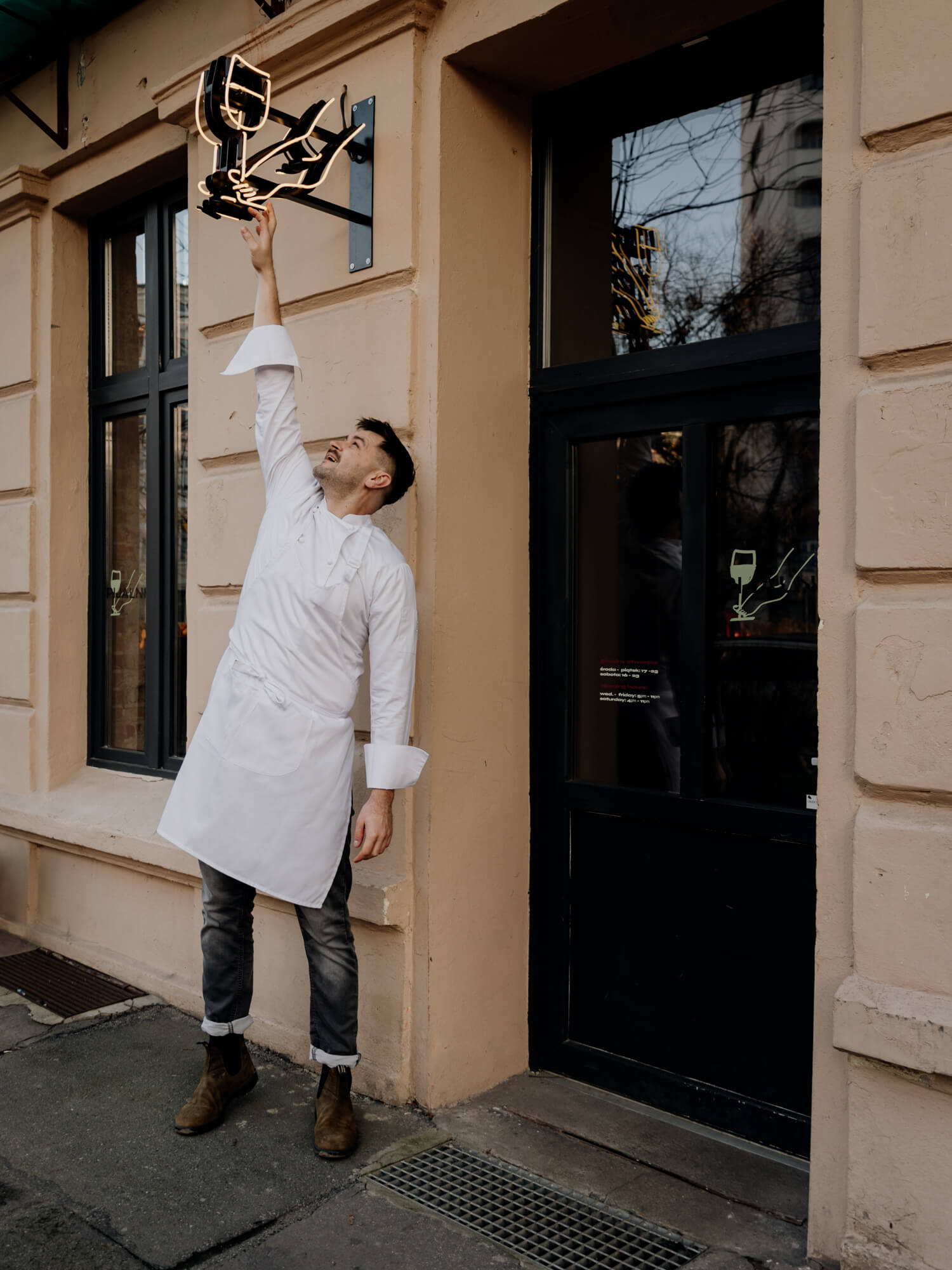 A person in a white outfit stands outside a building, raising their hand towards a sign above the door.