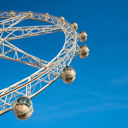 Partial view of a Ferris wheel with white framework and enclosed passenger pods against a clear blue sky.
