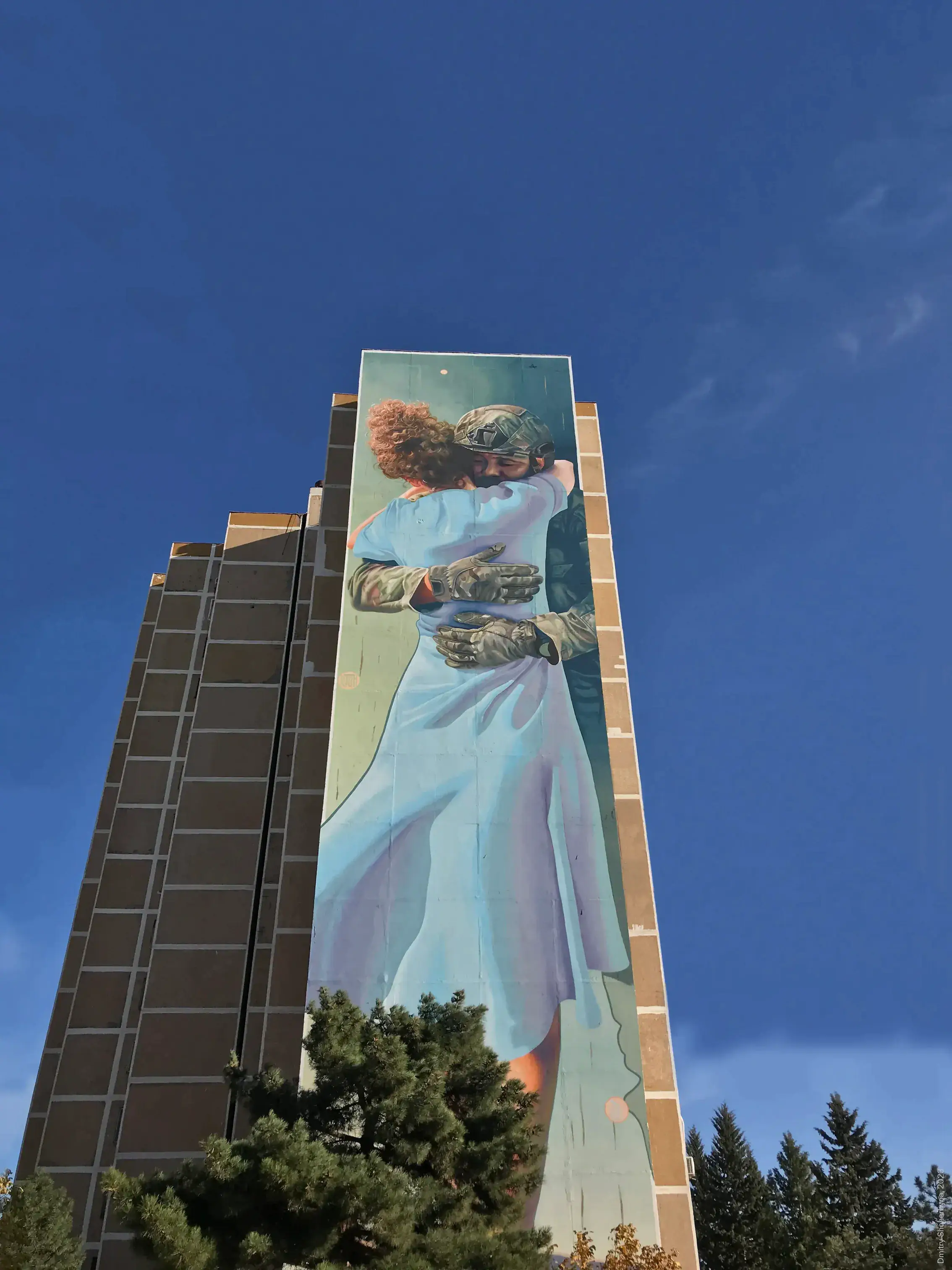 A tall mural on a high-rise building shows a soldier in combat gear and a helmet tenderly embracing a woman in a flowing light blue dress against a muted teal background.