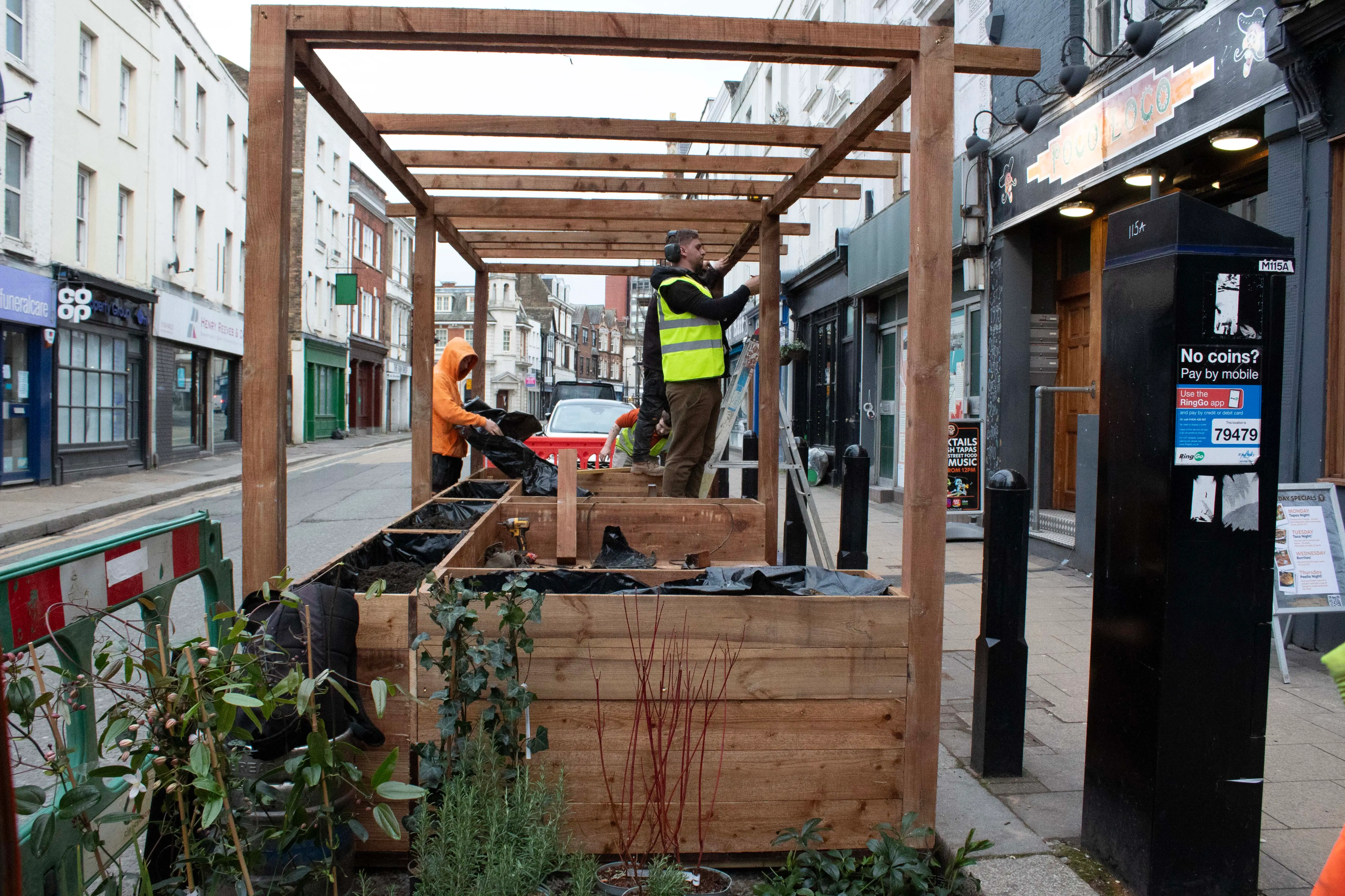 Construction area featuring wooden framework and workers, with greenery and nearby shops visible.