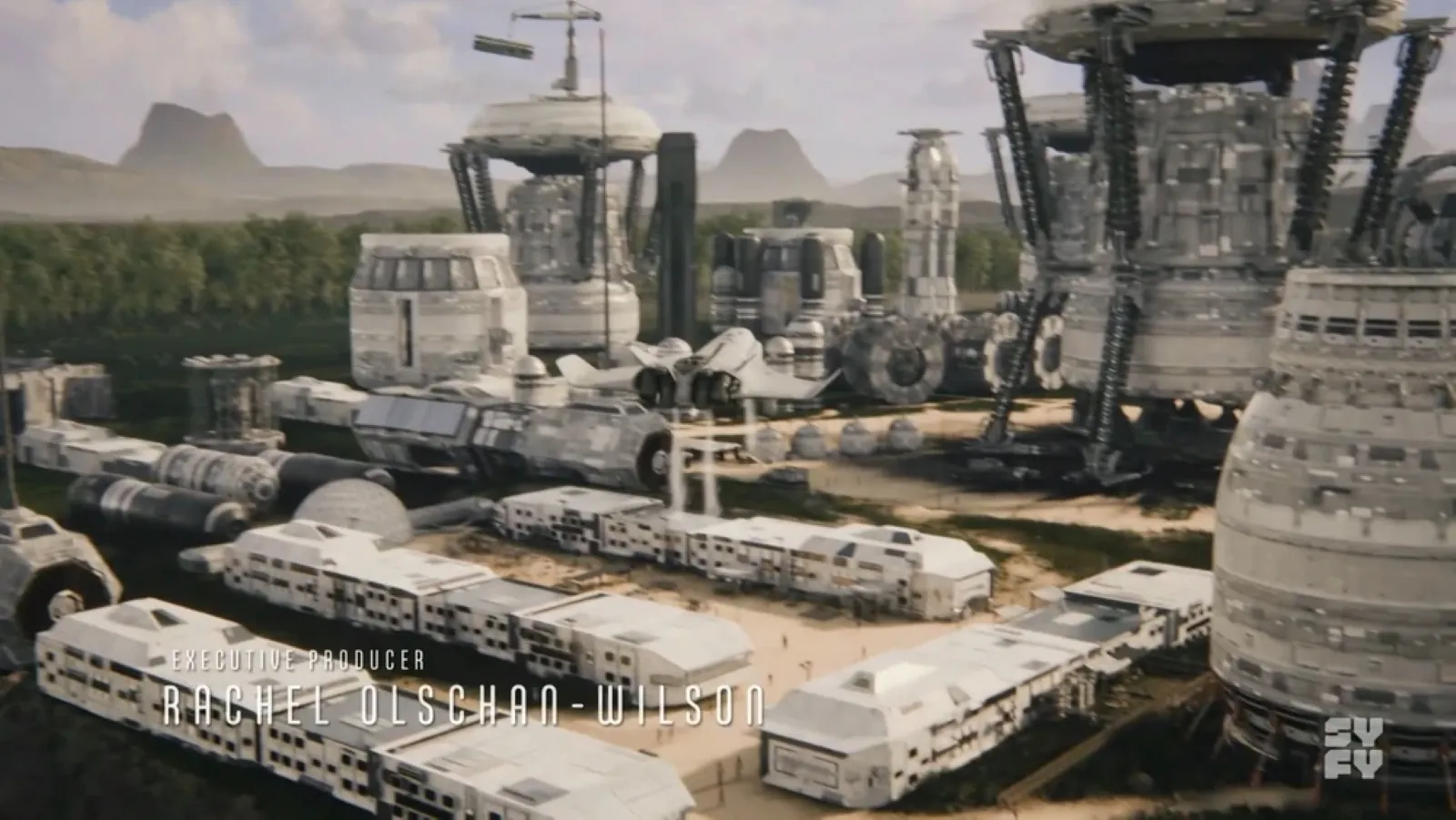 A futuristic space colony featuring large, technologically advanced structures and vehicles is set against a backdrop of lush green forests and distant mountains.