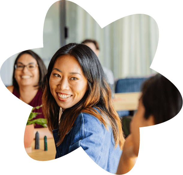 Smiling woman turning toward the camera while sitting at a table with coworkers in a casual meeting setting.