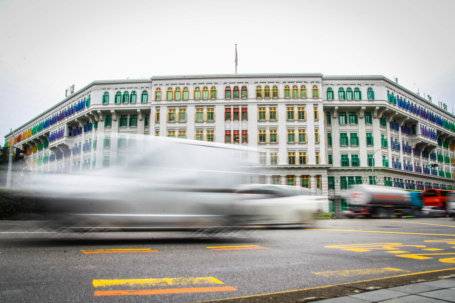 This image features the iconic Old Hill Street Police Station in Singapore. The building is well-known for its vibrant, multi-colored windows arranged in a rainbow pattern across its facade. The architecture is neoclassical and colonial in style.  In the foreground, there is a motion-blurred effect of fast-moving vehicles, adding a dynamic and modern contrast to the historical building. The street markings and vehicles suggest a busy urban setting, while the bright colors of the windows make the building visually striking and culturally distinctive.