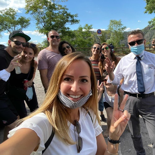 Group of people smiling outdoors, one wearing a face mask and another with a lowered mask, posing for a photo on a sunny day.