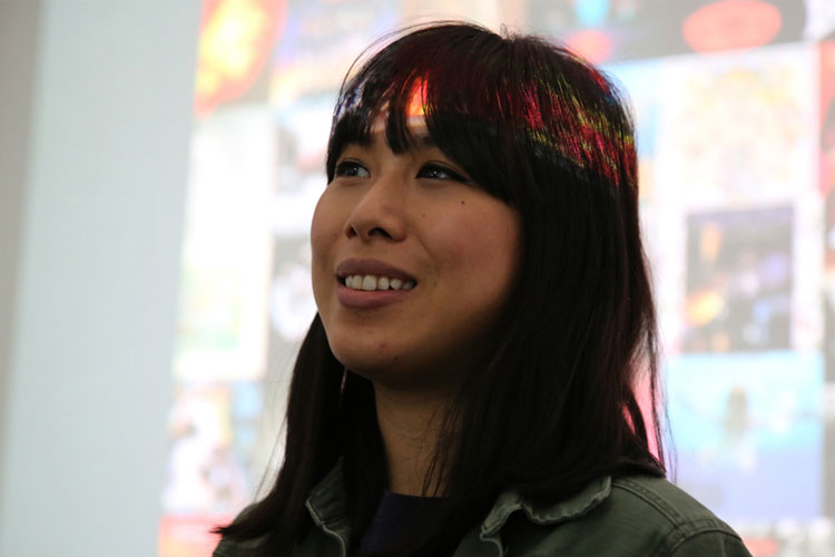 A woman with long dark hair and bangs smiles slightly while looking to her left. She wears a dark olive green jacket over a dark purple shirt. A colorful, rainbow-like streak is visible across her hair and across her forehead. The background is out of focus and includes blurred circular shapes.