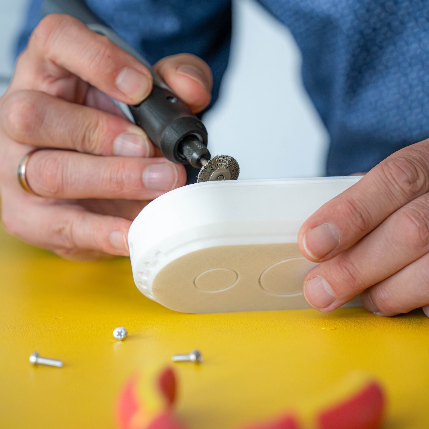 Hände bearbeiten mit einem rotierenden Werkzeug einen weißen 3D-gedruckten Prototyp auf einem gelben Untergrund