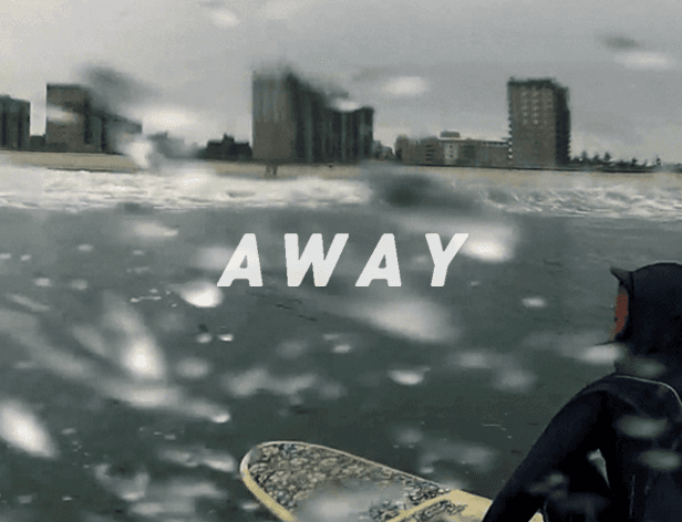 A person in a black wetsuit and hood carries a bright blue surfboard under their arm while walking across a snow-covered surface, with a tall building and structures in the background under a bright, overcast sky.