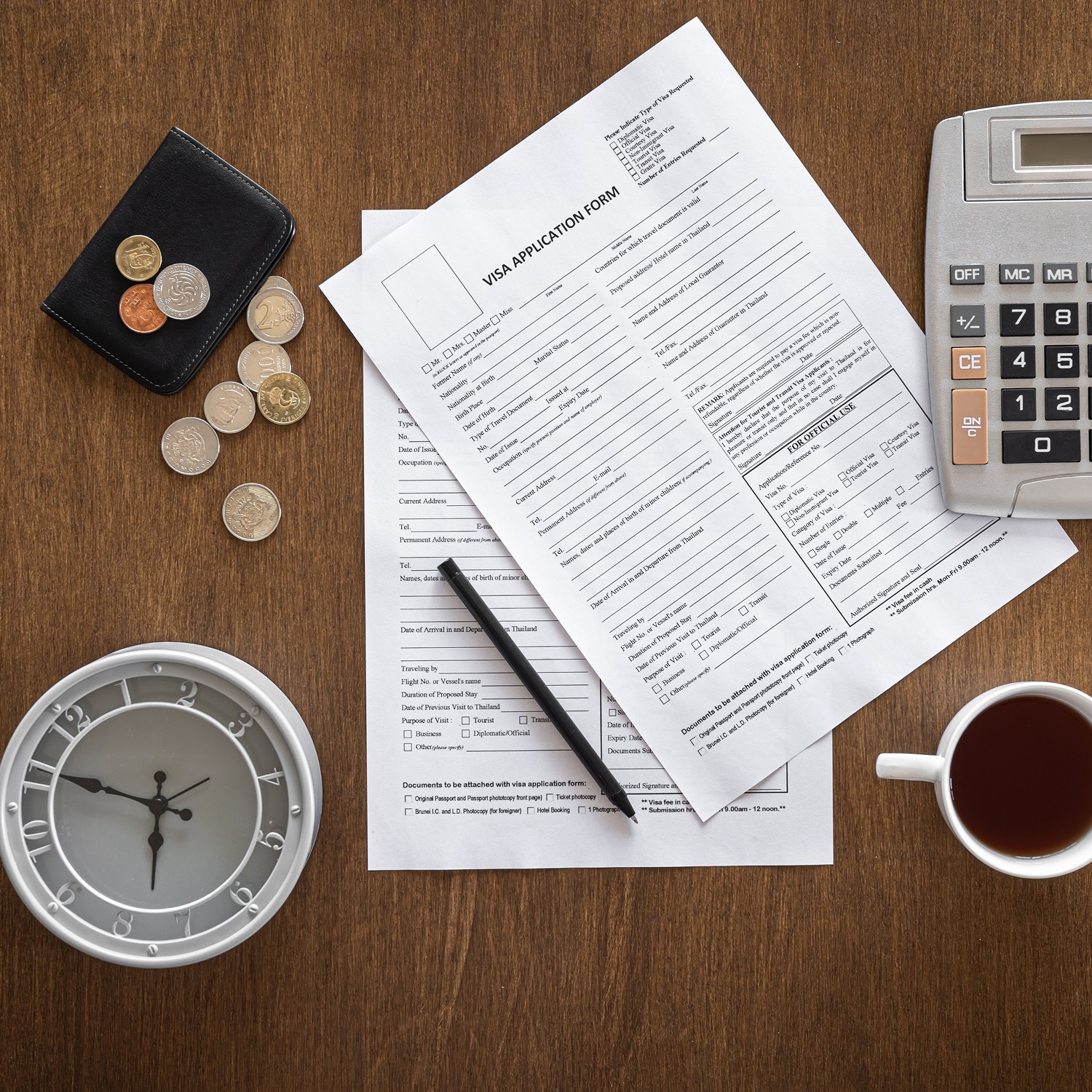 A visa application form lies on a wooden desk alongside a pen, calculator, coins, wallet, clock, and a cup of coffee, illustrating a workspace focused on financial planning and paperwork organization.
