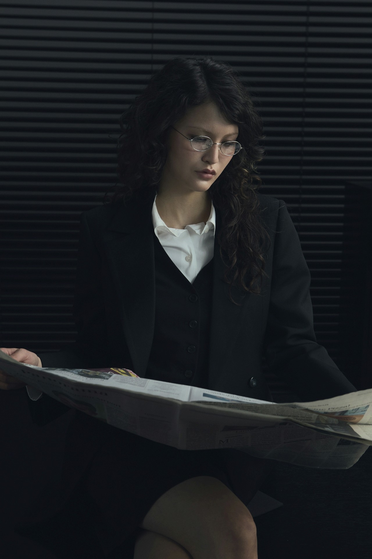 A woman in formal wear reading a newspaper while seated indoors.