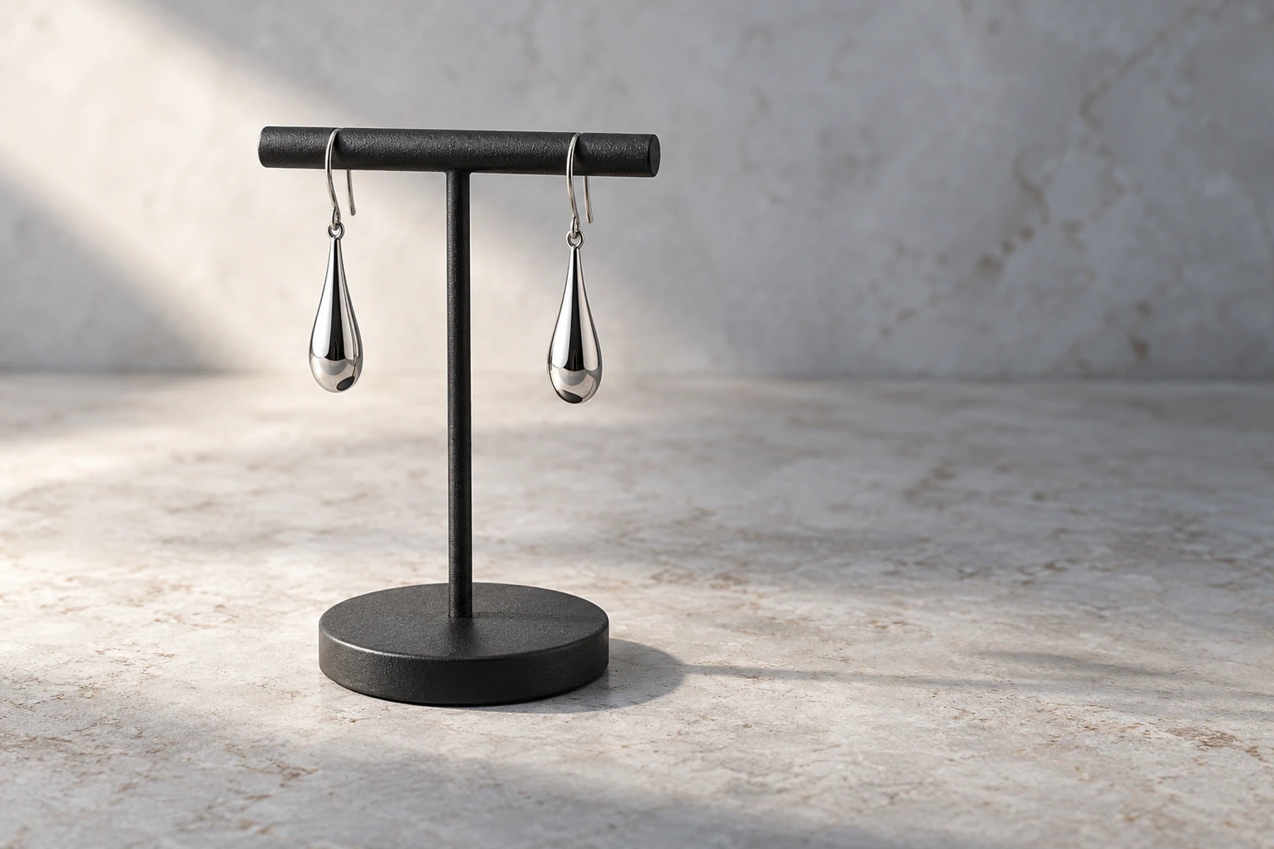 Conceptual silver teardrop earrings hanging from a charcoal T-bar above a pale stone surface