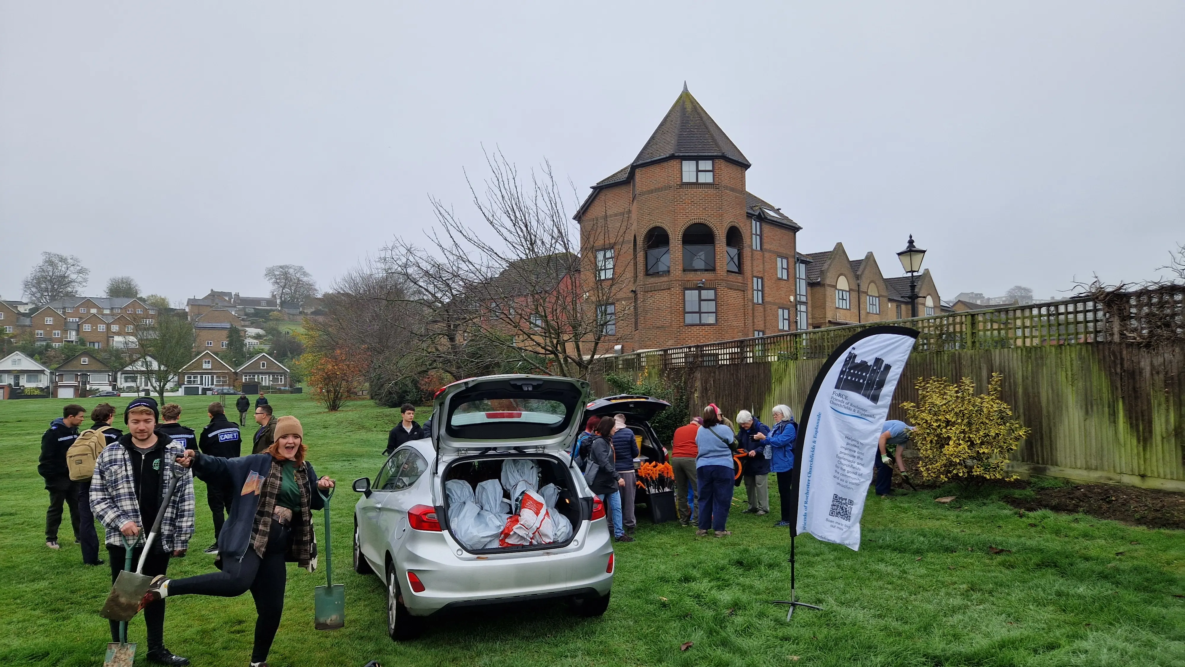 A group of people gathers near a car and a building, with a banner displayed in a grassy area.