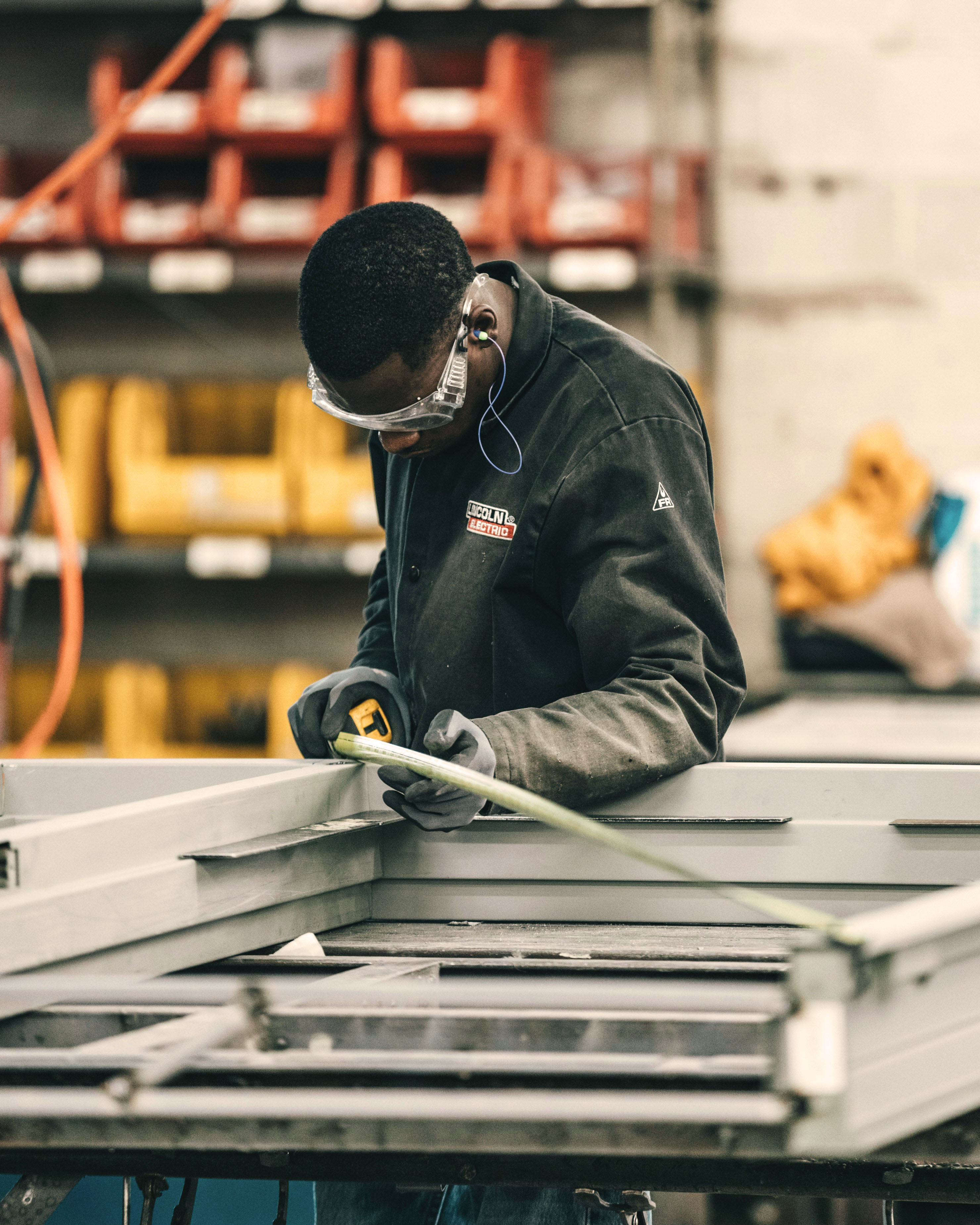 A person in work attire focuses on assembling metal components in a workshop filled with tools and materials.