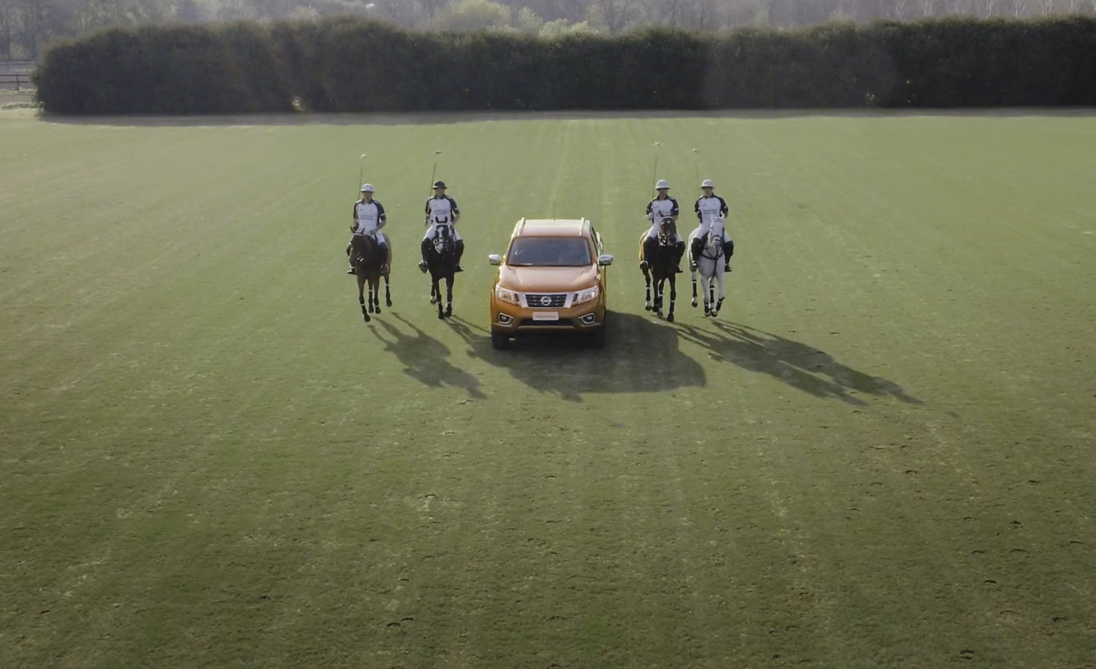 A bright orange SUV is flanked by four polo players on horseback, positioned symmetrically on a vast green field, exuding a dynamic and sporty vibe.