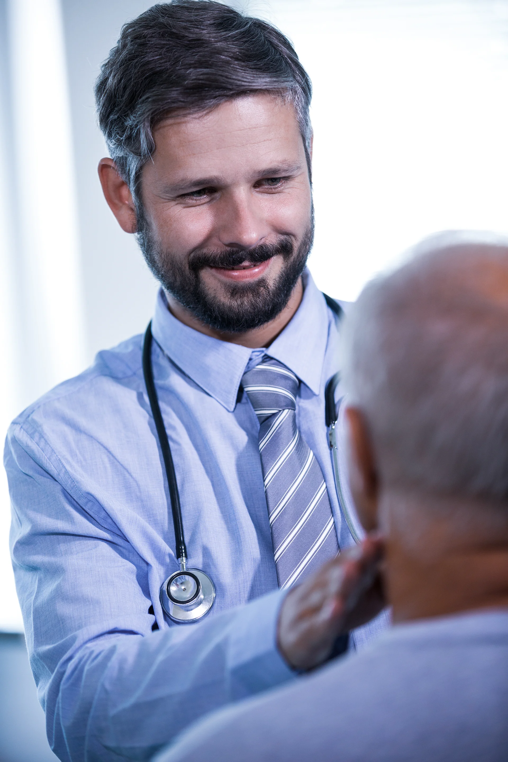 Um médico sorridente com barba e estetoscópio examina um paciente idoso em um consultório médico iluminado, examinando o pescoço dele.