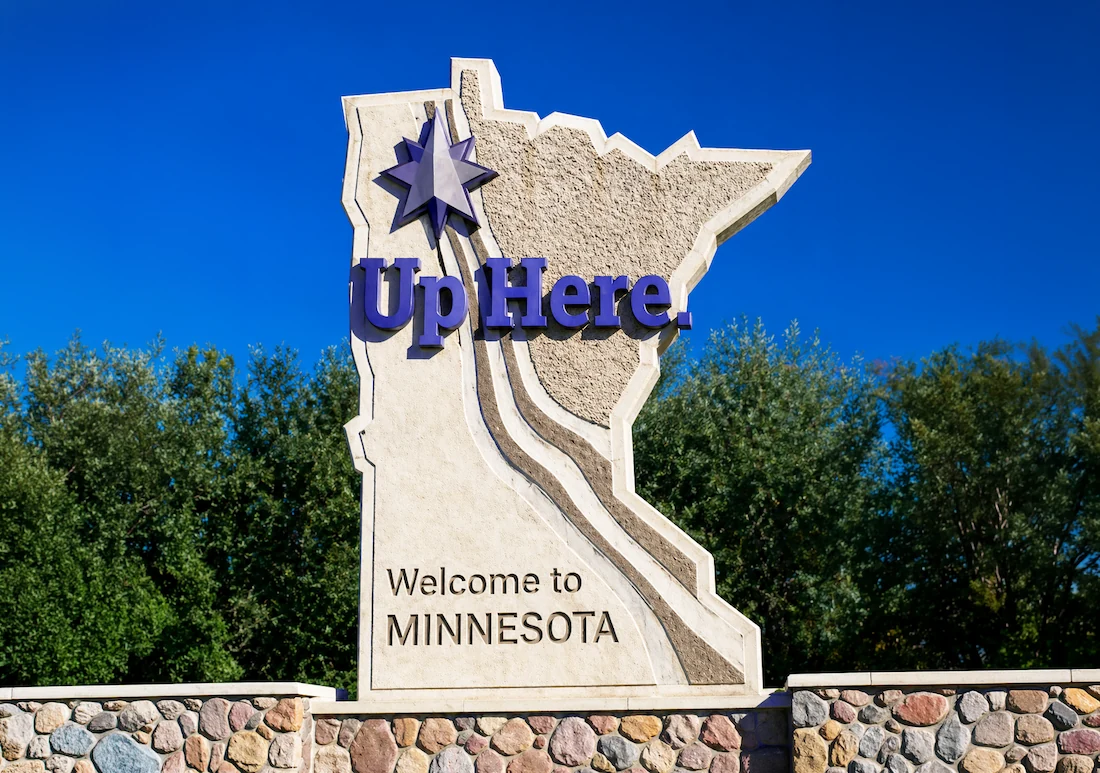 The redesigned Minnesota state welcome sign monument featuring the new eight-pointed star mark and Up Here. in raised three-dimensional navy lettering, with Welcome to Minnesota carved into the stone below, set against a deep blue sky.
