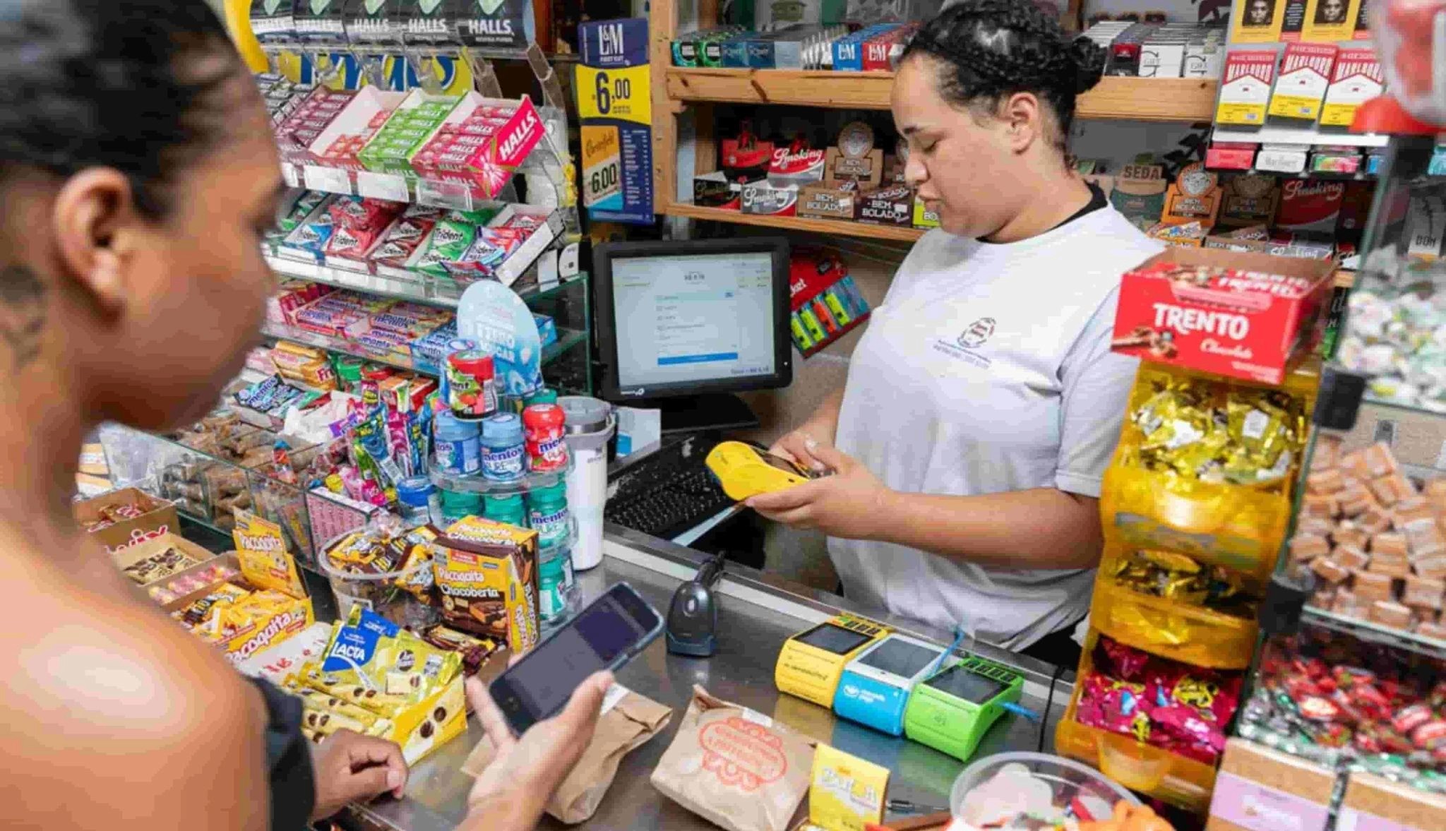 Atendente de loja de conveniência finaliza venda no PDV usando maquininha de cartão, enquanto cliente segura o celular. O balcão está cheio de doces e produtos.
