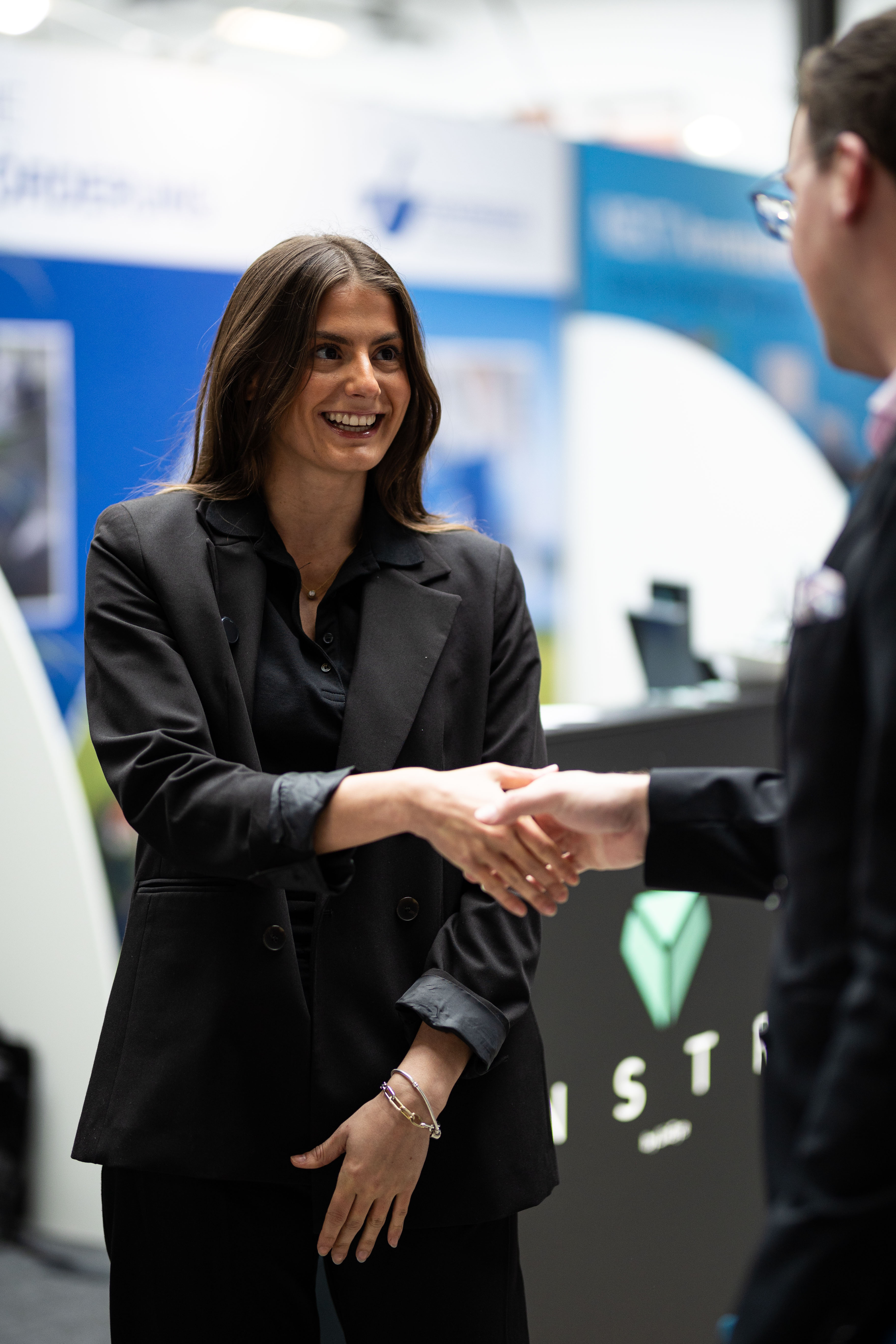 Eine junge Frau in einem schwarzen Business-Anzug lächelt, während sie einem Mann (dessen Rücken und rechte Seite im Vordergrund sichtbar sind) die Hand schüttelt. Die Szene spielt auf einer Messe oder Konferenz, erkennbar an einem Messestand und verschiedenen Logos im leicht unscharfen Hintergrund, darunter ein stilisiertes, grünes Rauten-Logo mit der Beschriftung 'ISTR'.