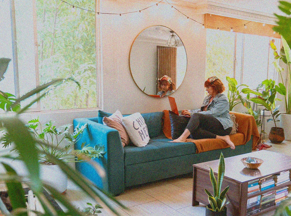 In a bright, sunny, plant-filled room, a barefoot therapists sits on blue couch with throw pillows, working on her laptop