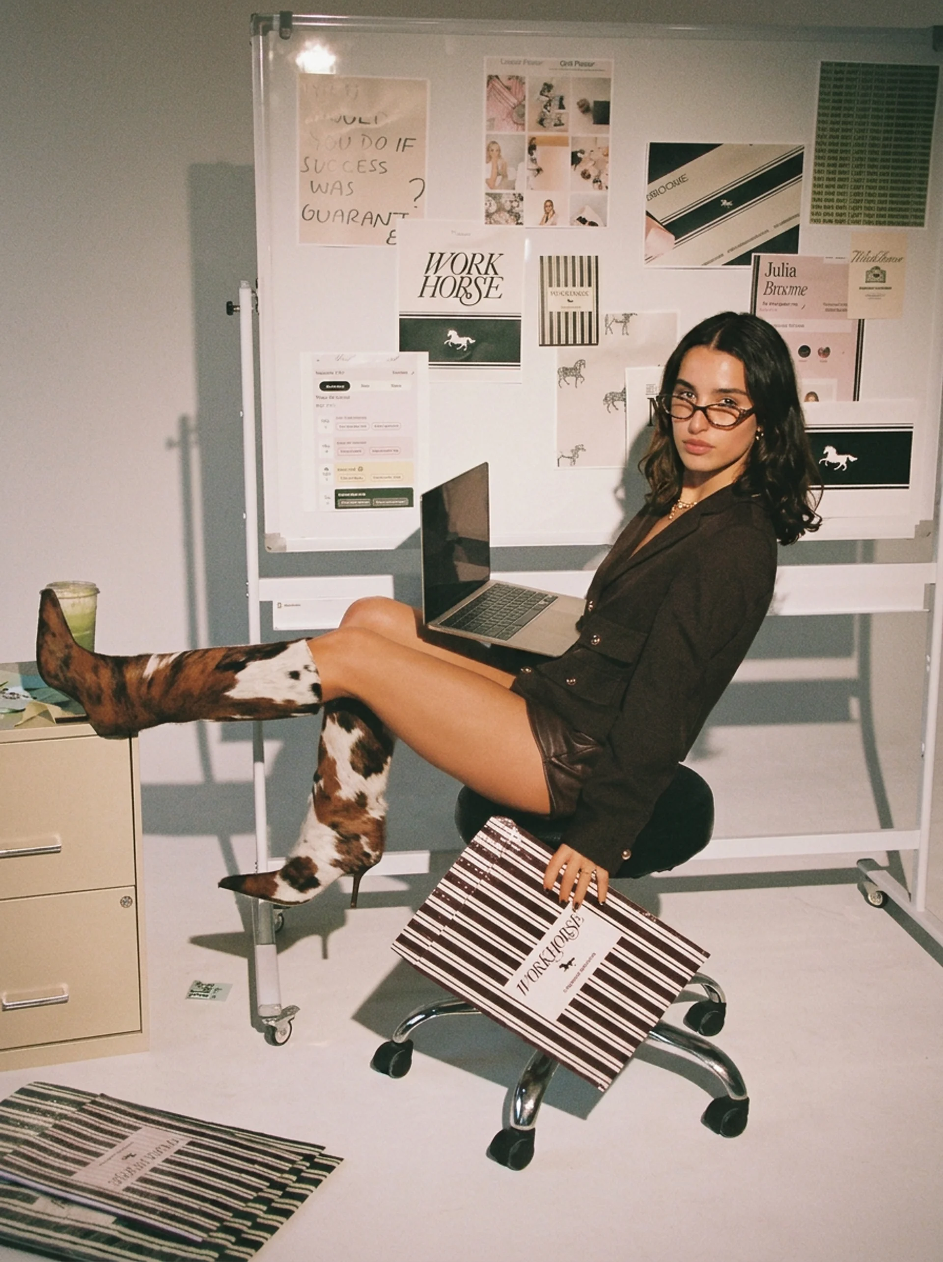 Amelia in cow-print boots, perched on a stool with a laptop in front of a moodboard