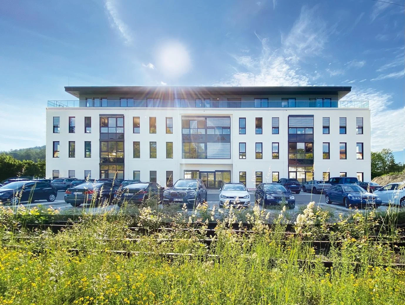 Modernes Bürogebäude in Weiß mit dunklem Dach und vielen Fenstern, davor ein Parkplatz mit mehreren Autos unter blauem Himmel.