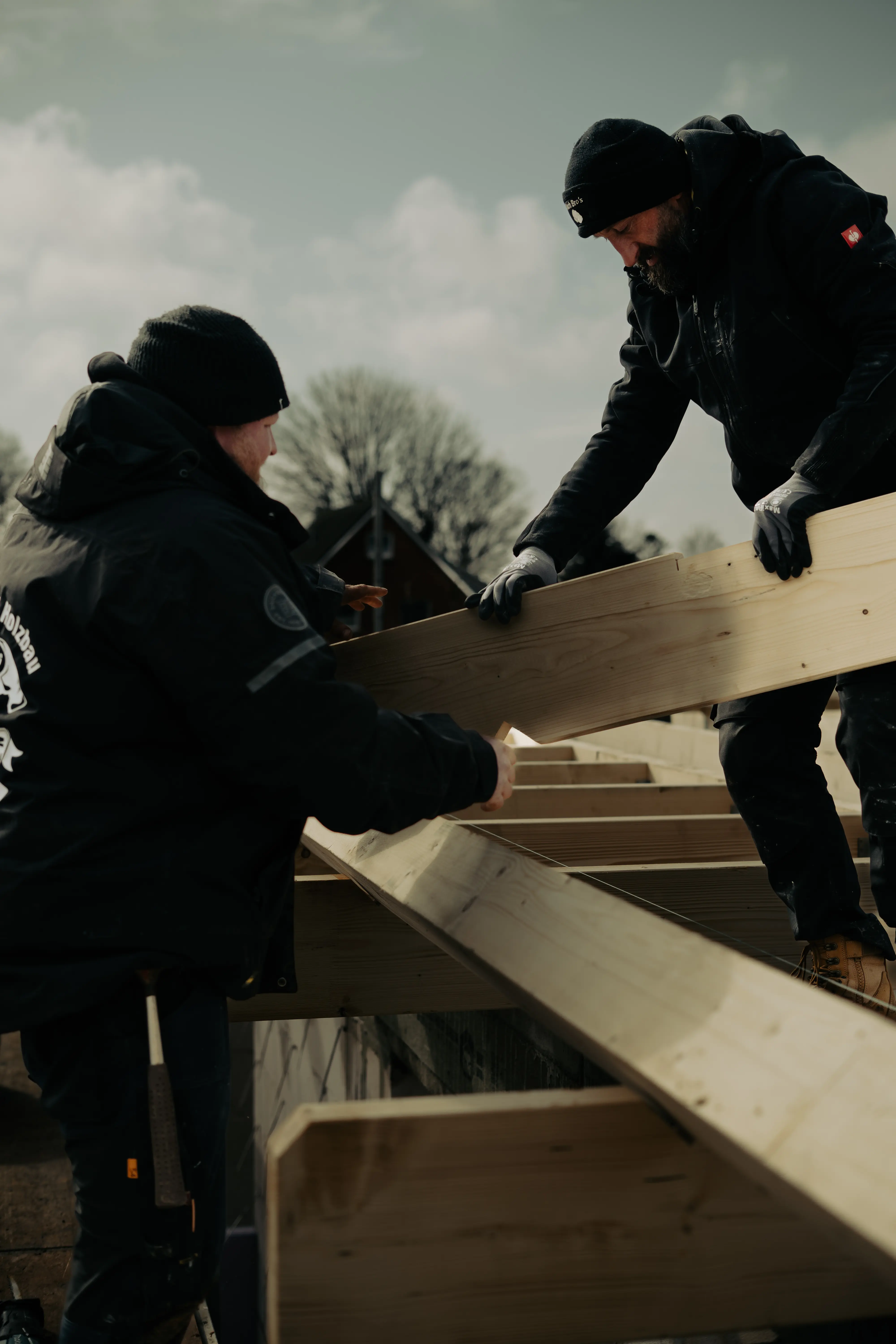 Zwei Bauarbeiter in schwarzer Winterkleidung errichten im Freien unter einem bewölkten Himmel eine Holzkonstruktion und konzentrieren sich dabei auf das Ausrichten großer Holzbalken.