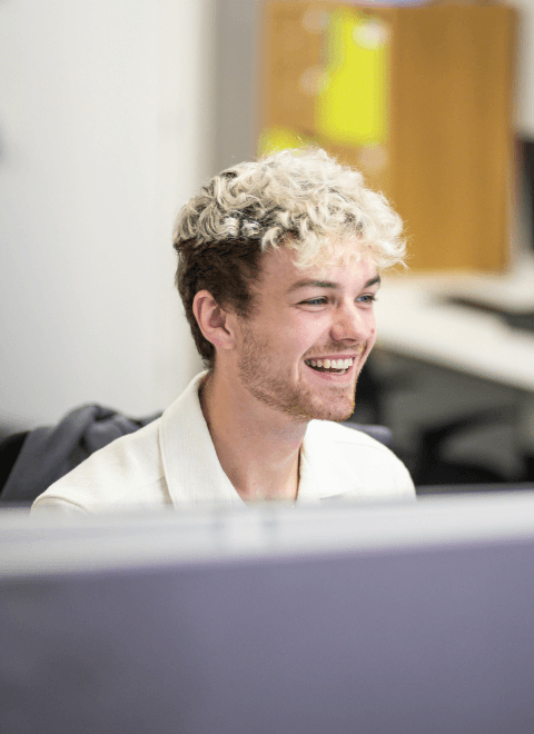 Male with blonde, curly hair smiling at his computer.