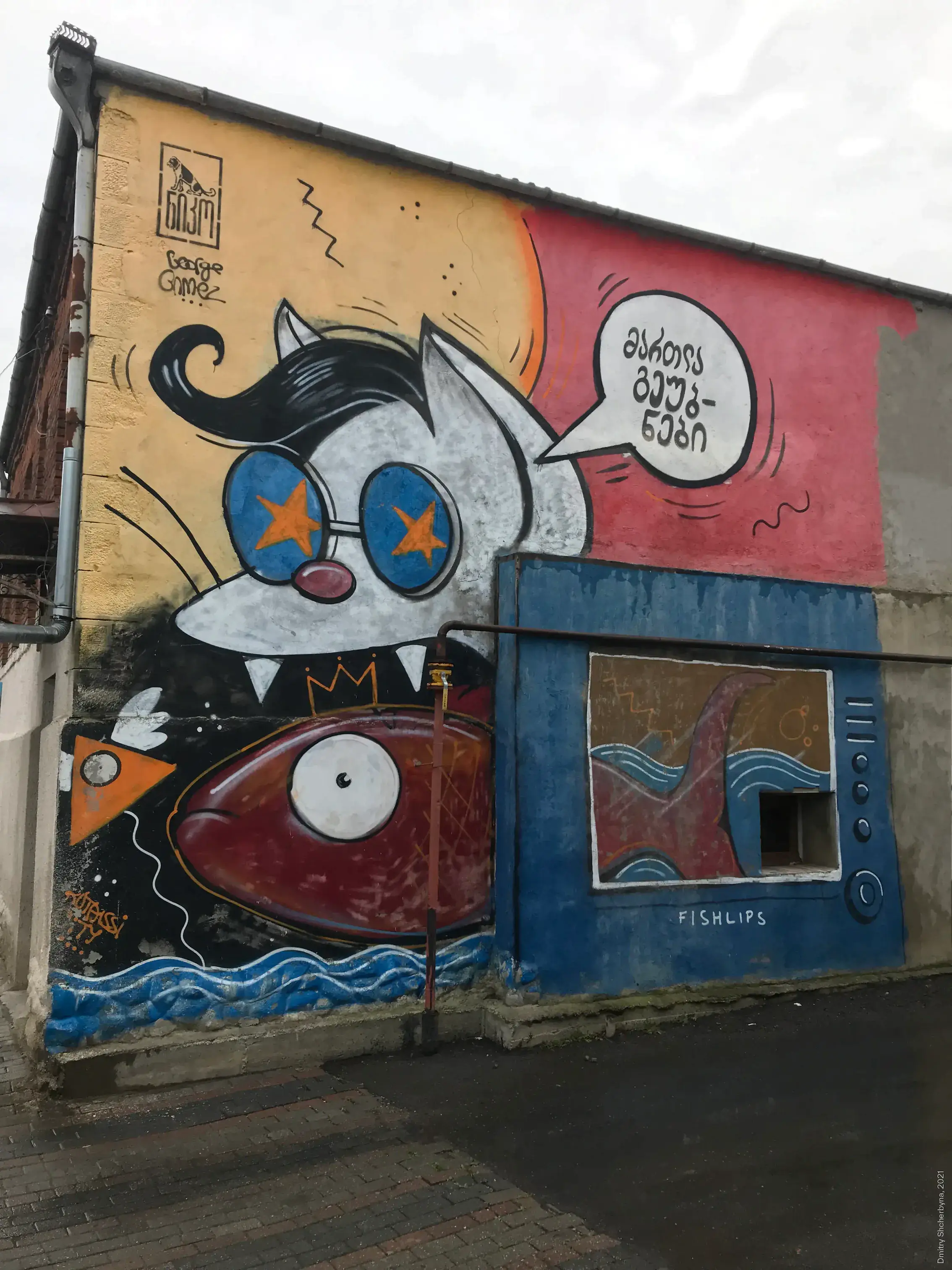 A playful mural featuring a wide-eyed cartoon cat wearing star-shaped glasses and sharp fangs, looming over a red fish crowned with a crown, next to a blue TV set labeled "FISH-LIPS" showing a fishtail.